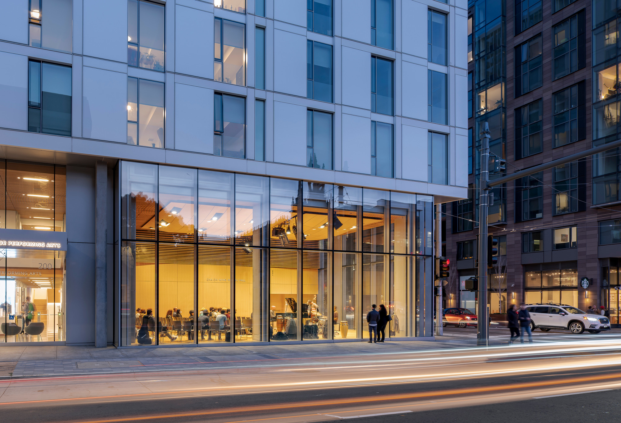 Ground-level view of the double-height glass lobby entrance with pedestrians passing at dusk