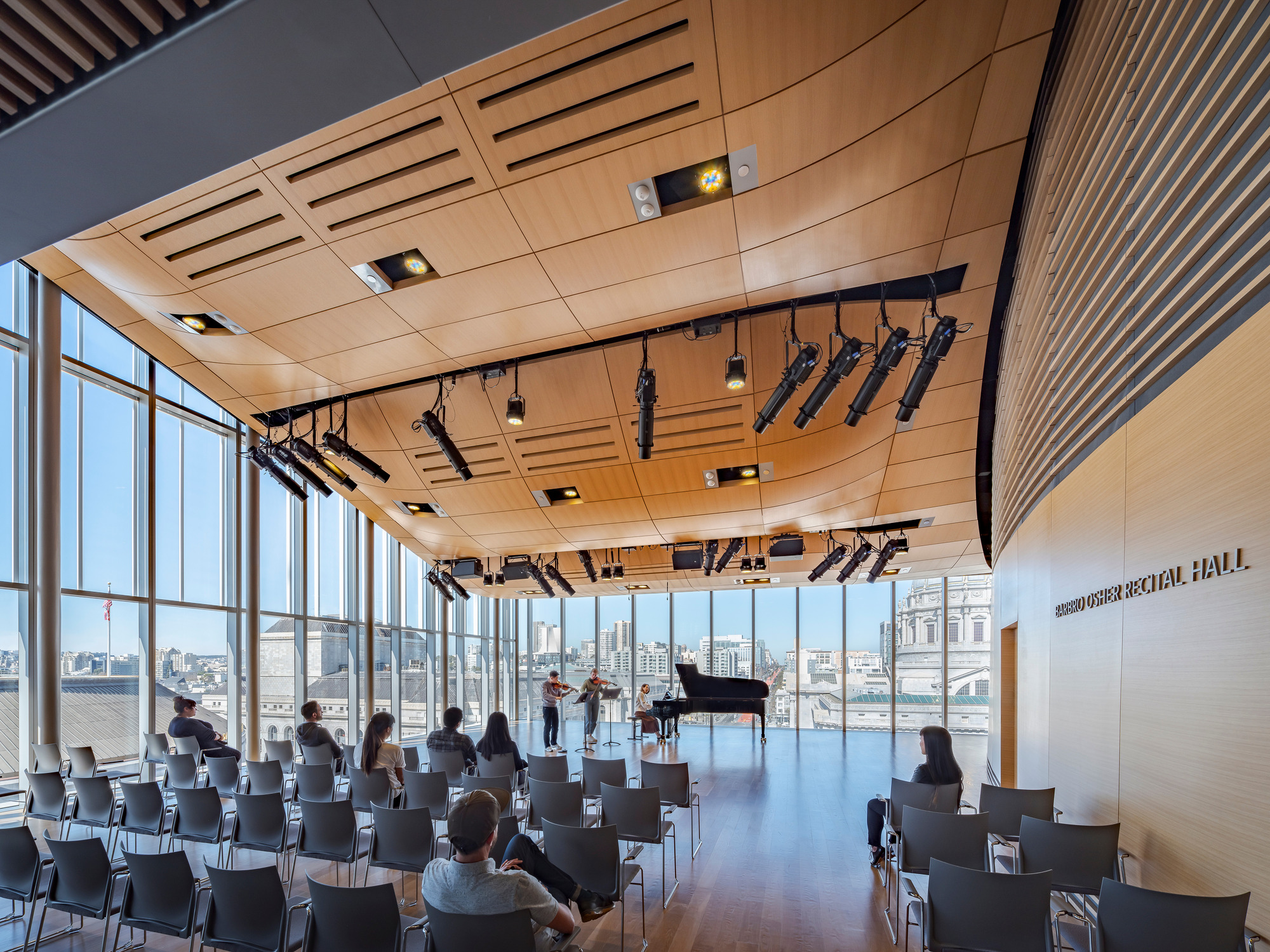 Recital hall with timber ceiling panels, floor-to-ceiling glazing, and audience seated facing a grand piano