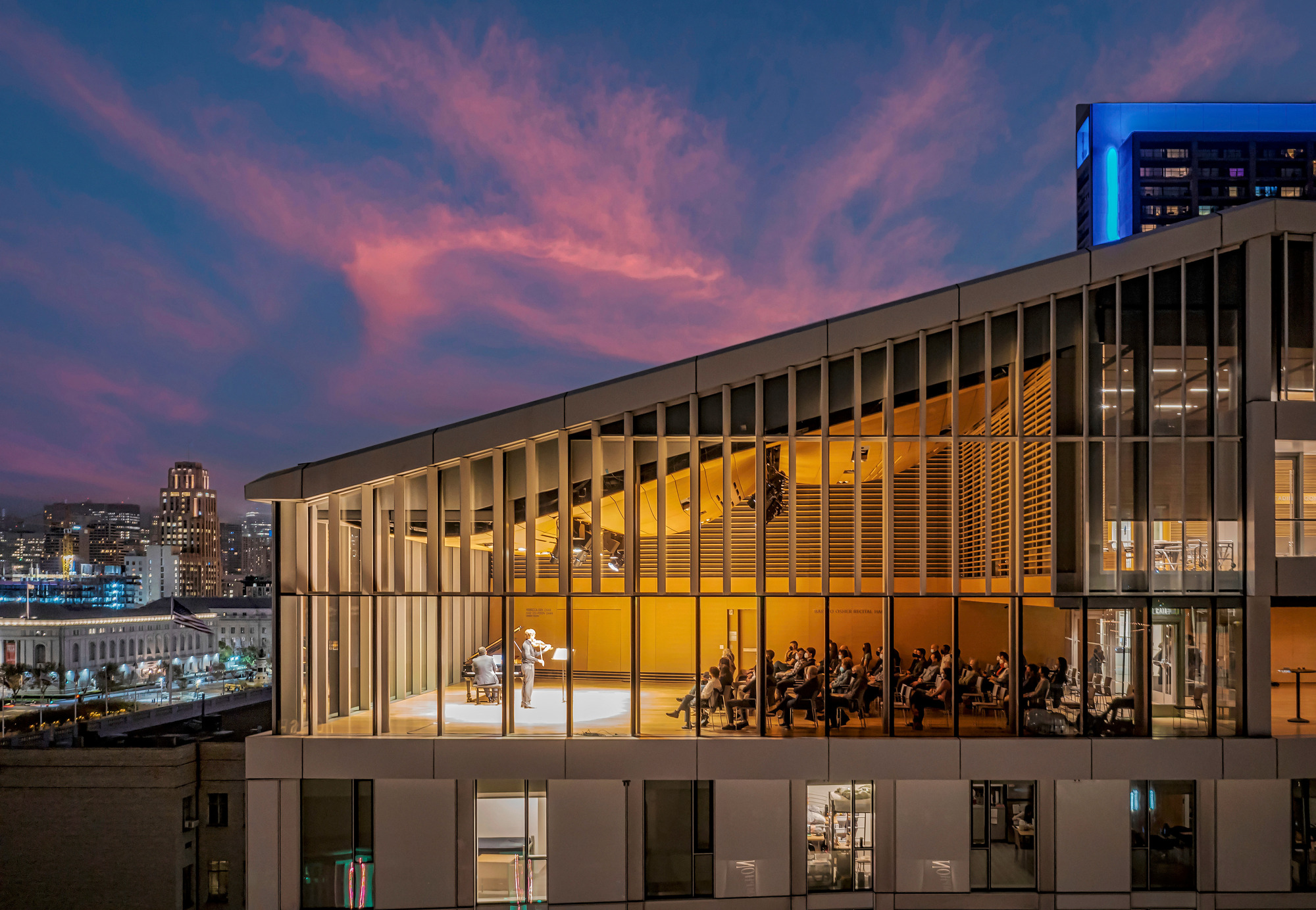 Glass-walled rehearsal room with vertical timber louvers and an orchestra visible inside under pink evening clouds