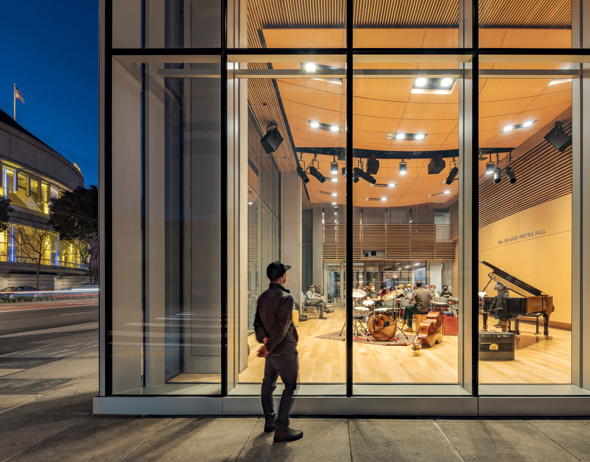 Glass storefront revealing a rehearsal room with timber ceiling and a passerby at dusk