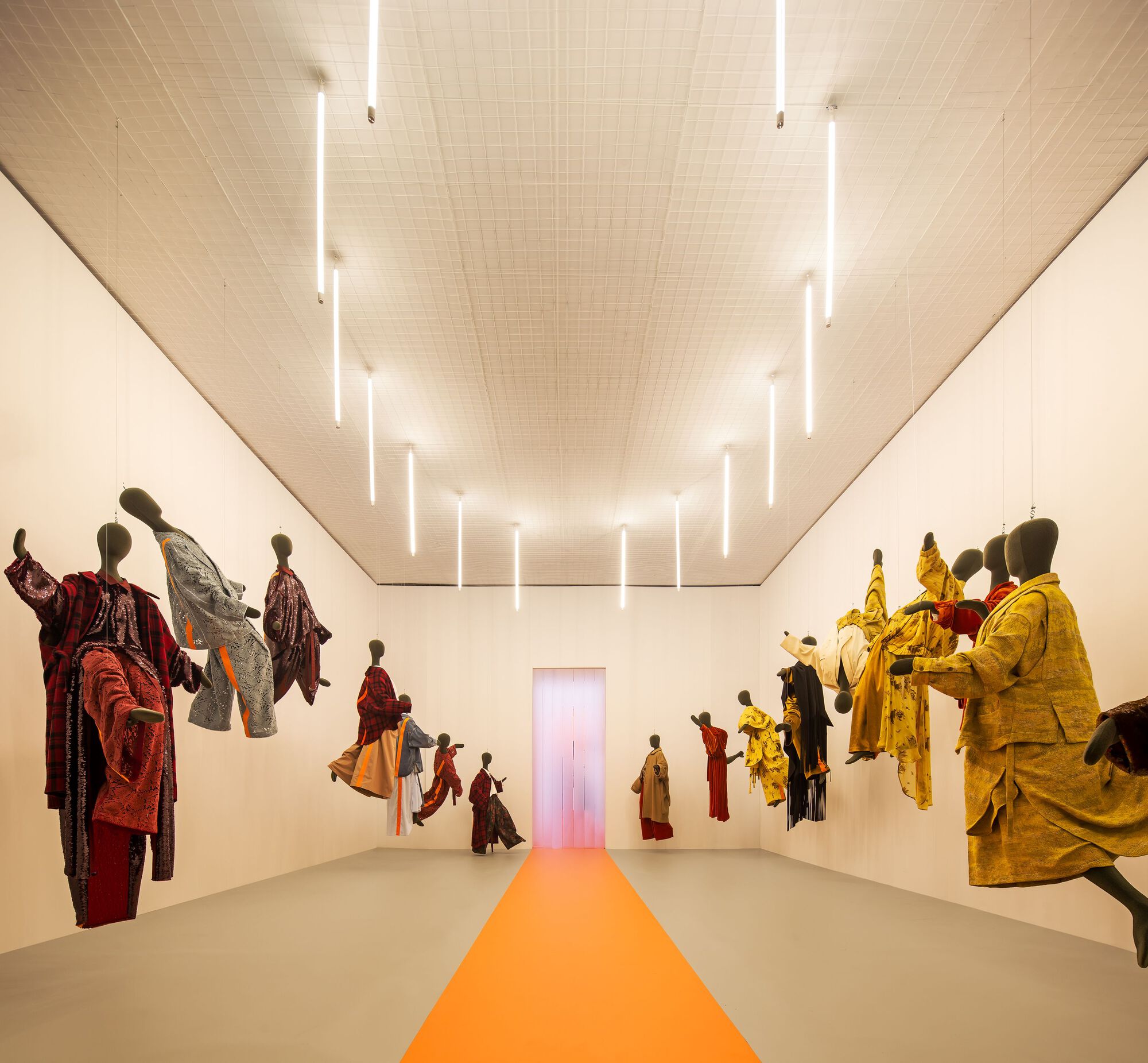 Gallery interior with suspended garments on mannequins and an orange floor stripe leading to a glowing doorway