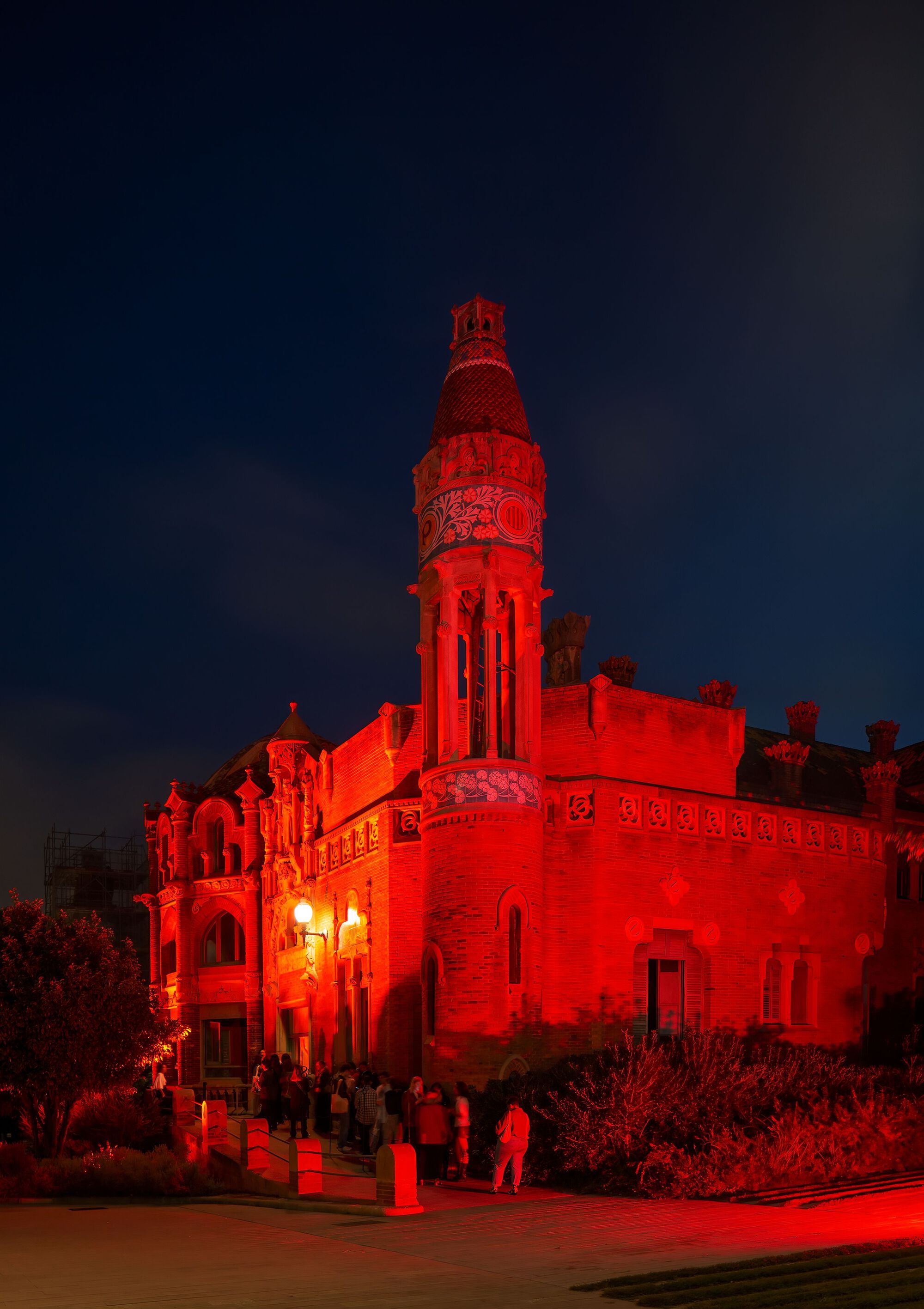 Ornate tower with carved detailing illuminated in red light at night with visitors gathered below