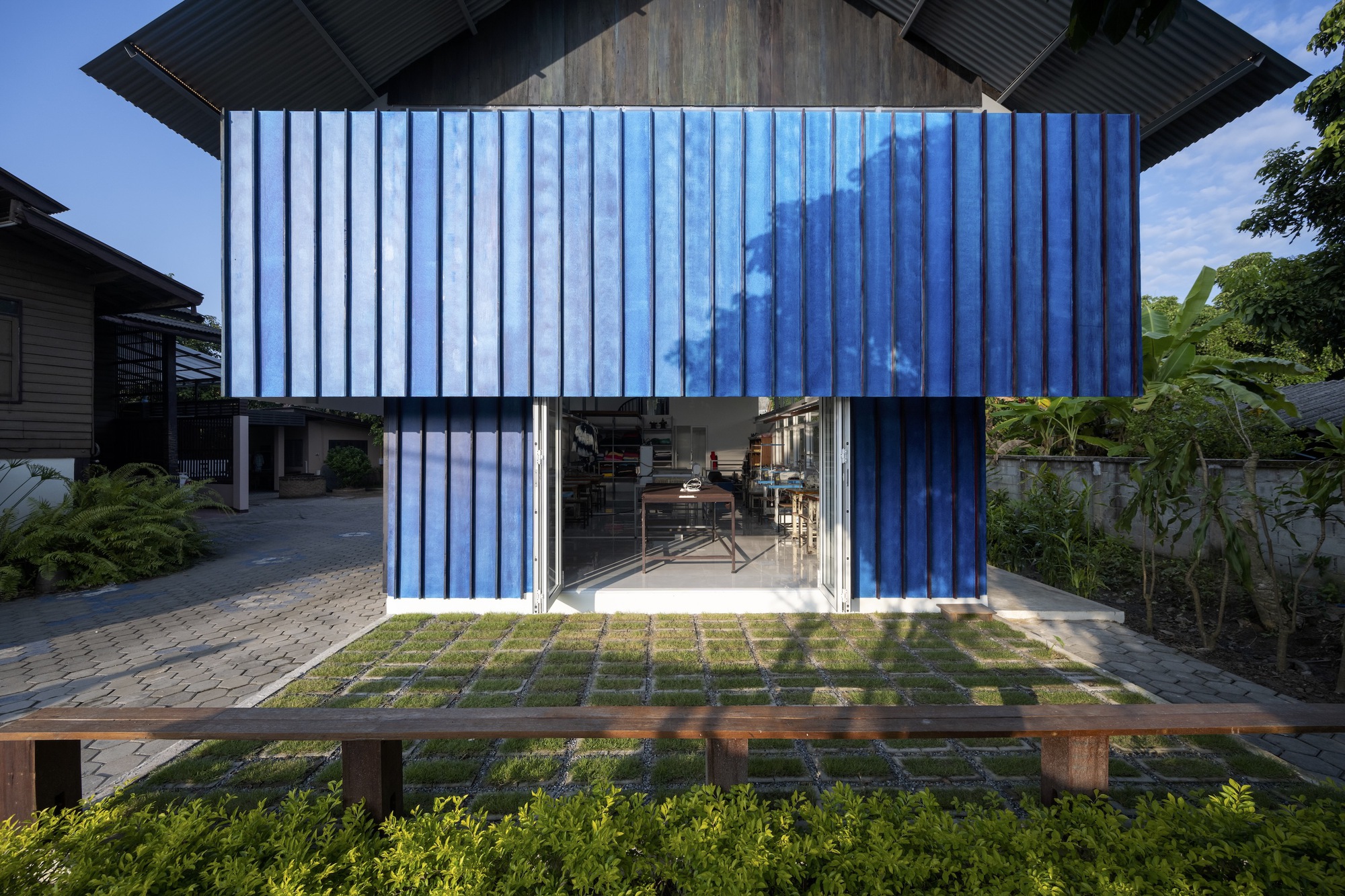 Blue corrugated metal volume with folding glass doors opening onto a gridded paver and grass courtyard