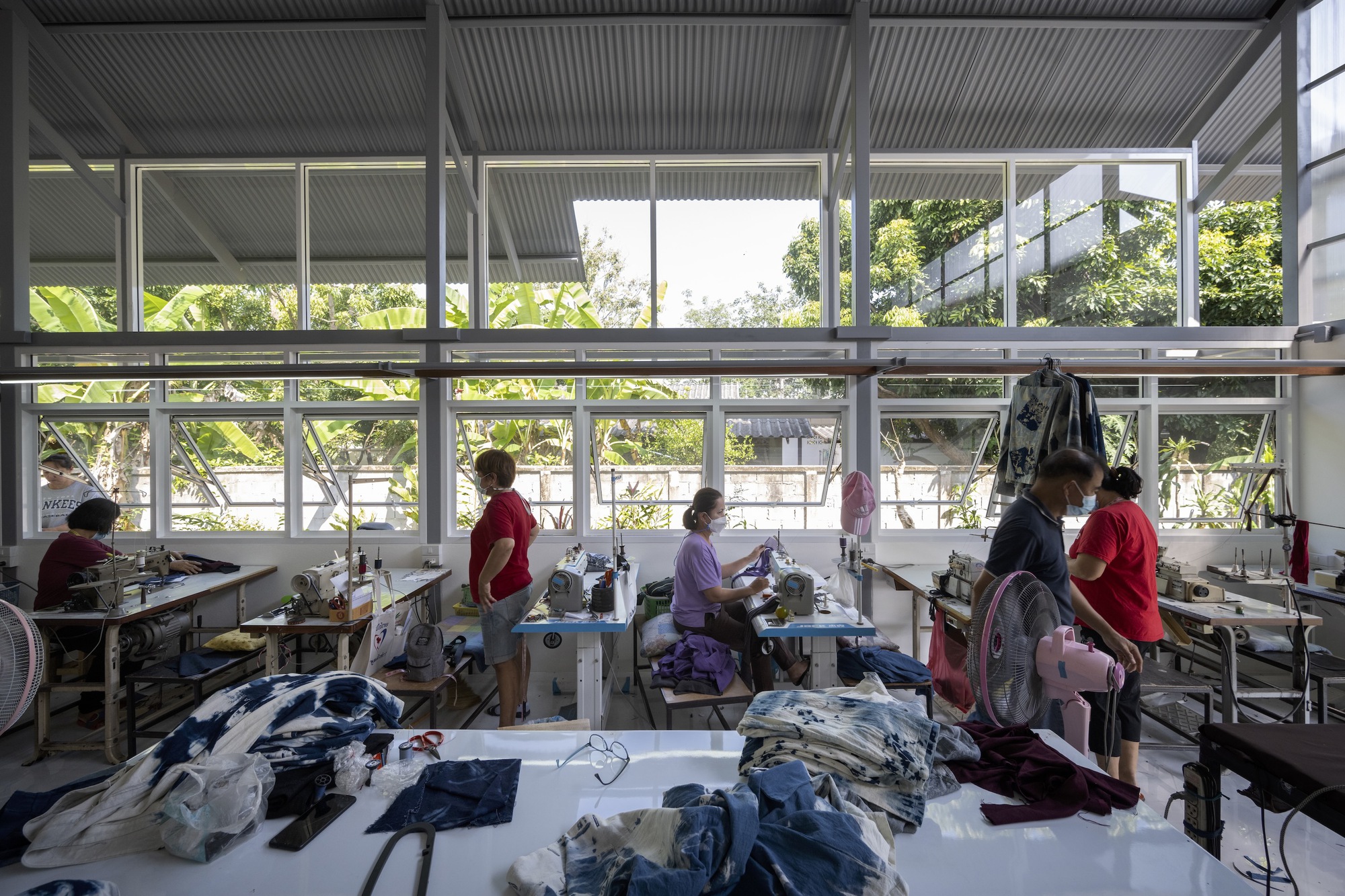 Workshop interior with workers at sewing stations beneath steel frame windows overlooking planted vegetation
