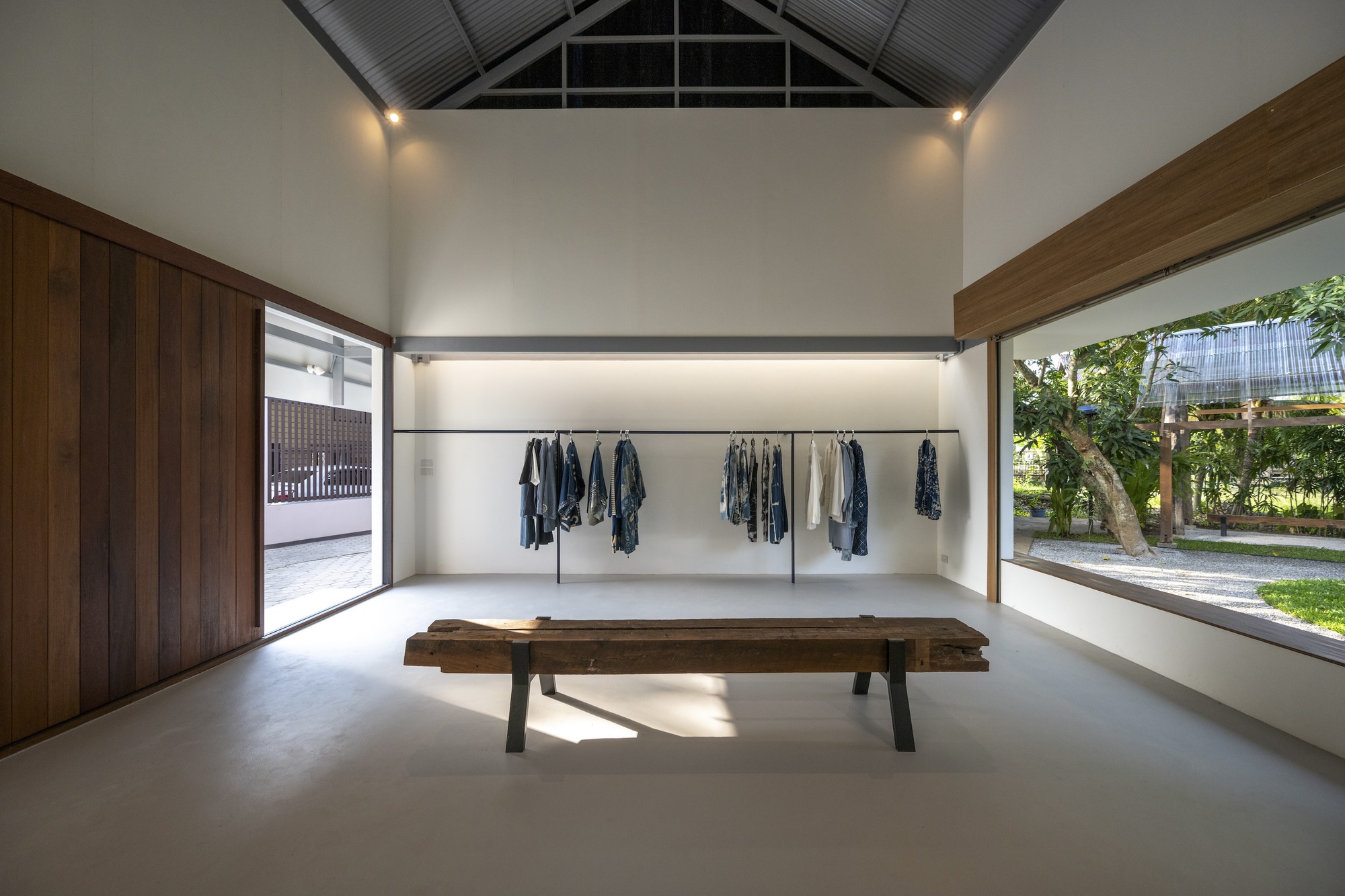 Retail space with timber bench, hanging garments, and horizontal windows framing the garden outside