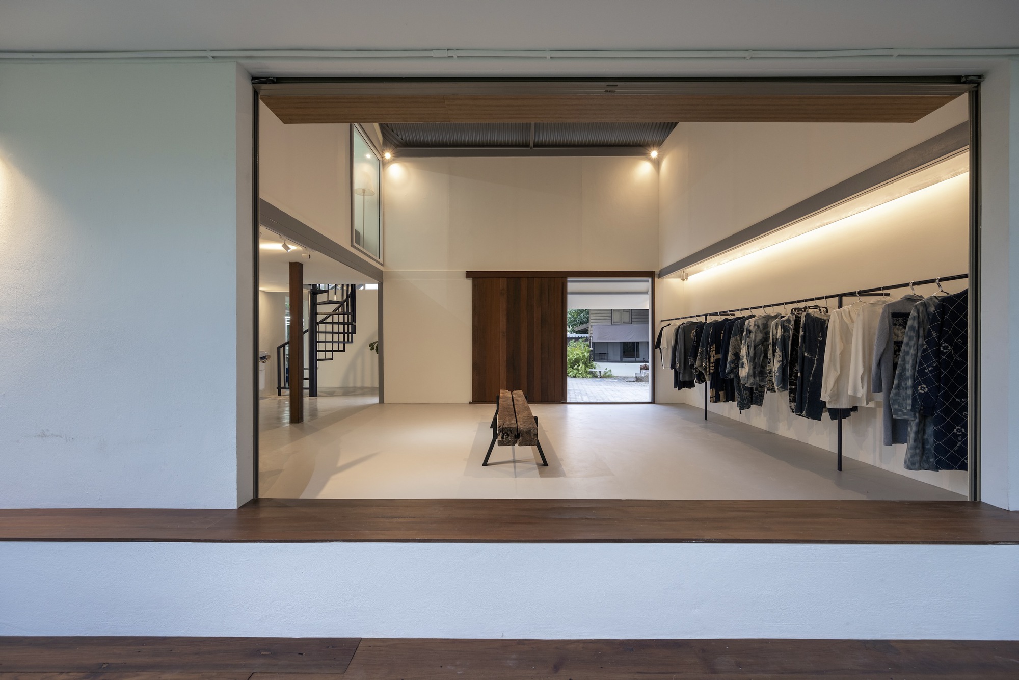 View through wide timber-framed opening showing the studio interior with clothing racks and spiral stair