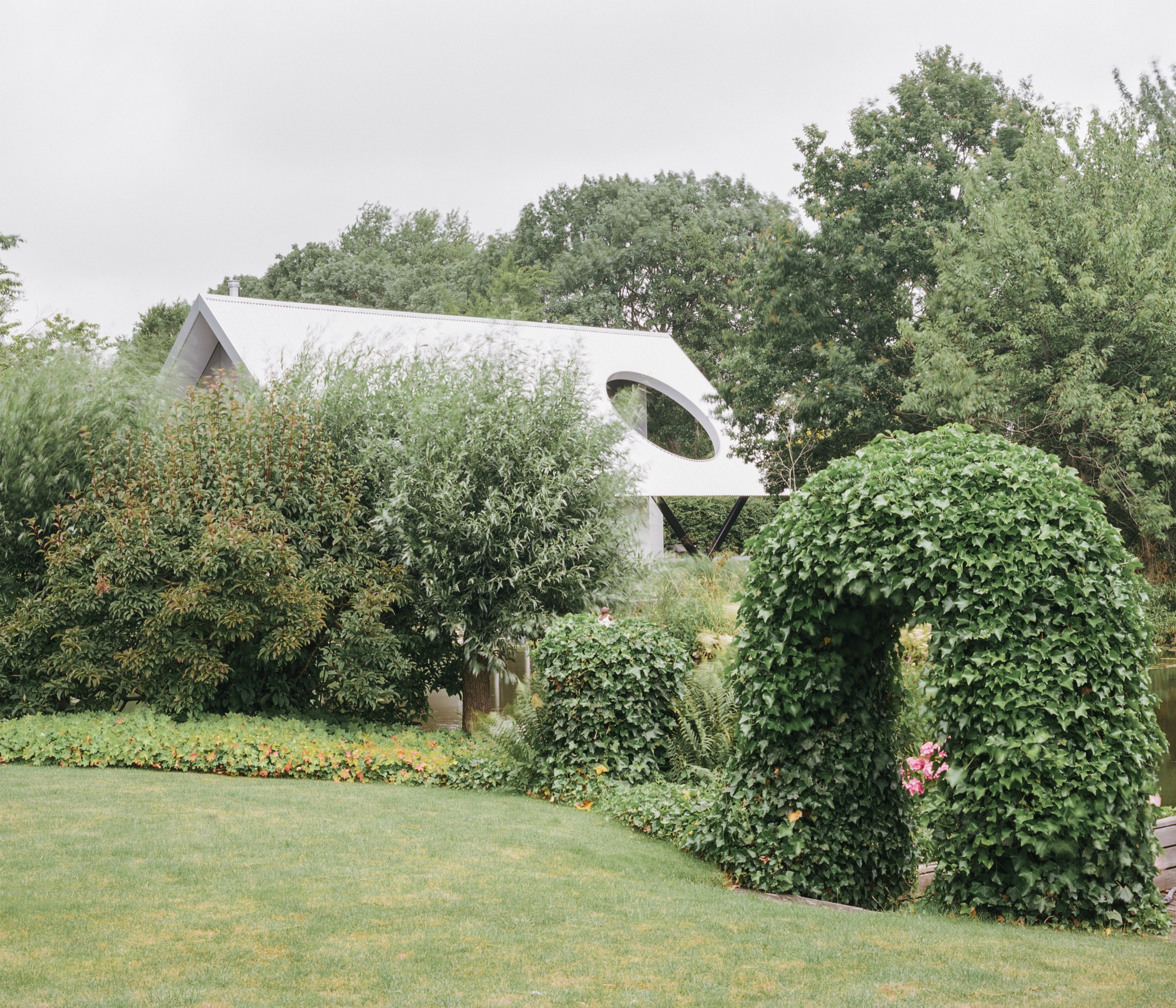 View through mature garden showing the corrugated roof and circular window beyond ivy-covered walls