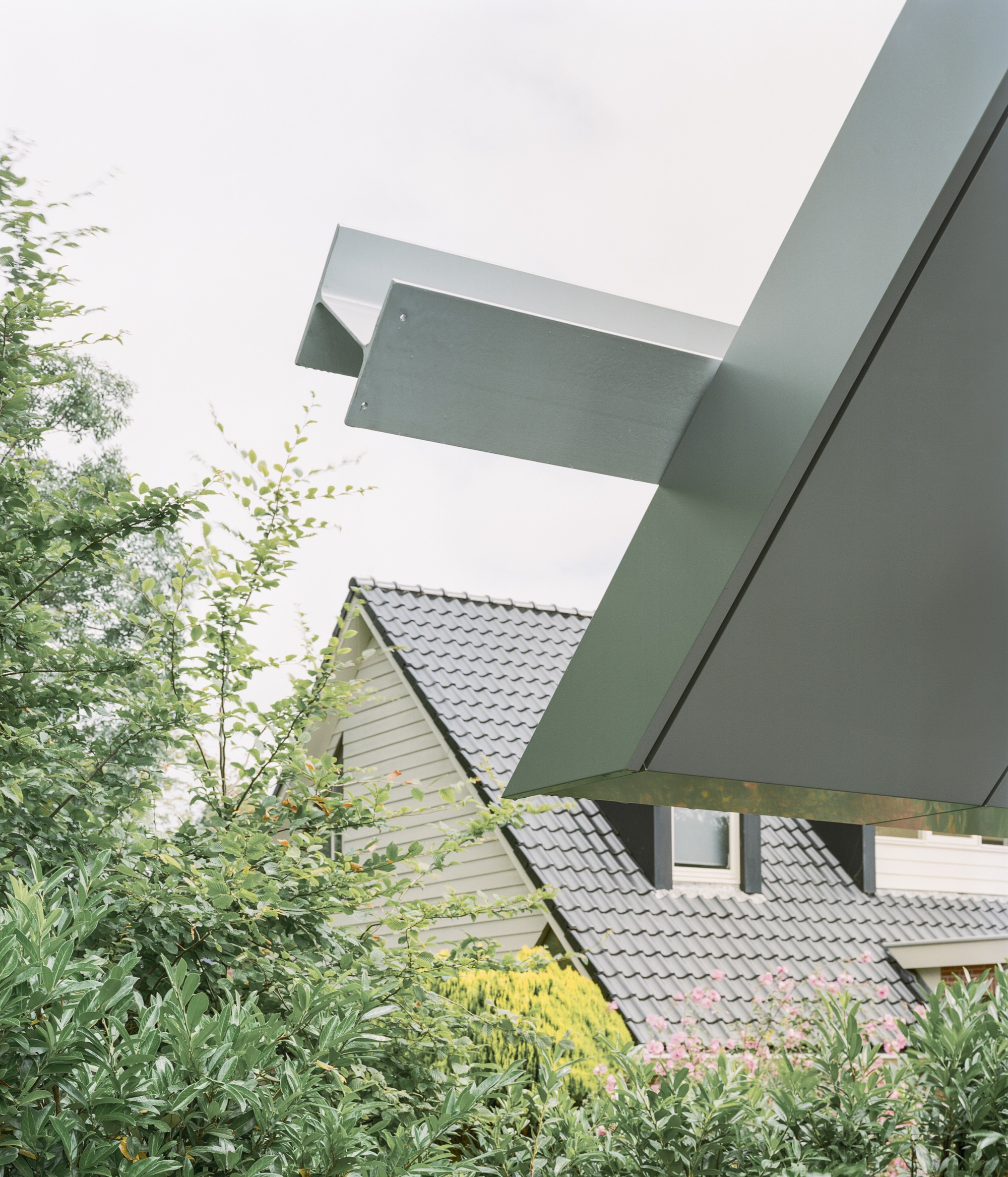 Cantilevered metal roof planes extending over neighboring tiled roof and garden plantings