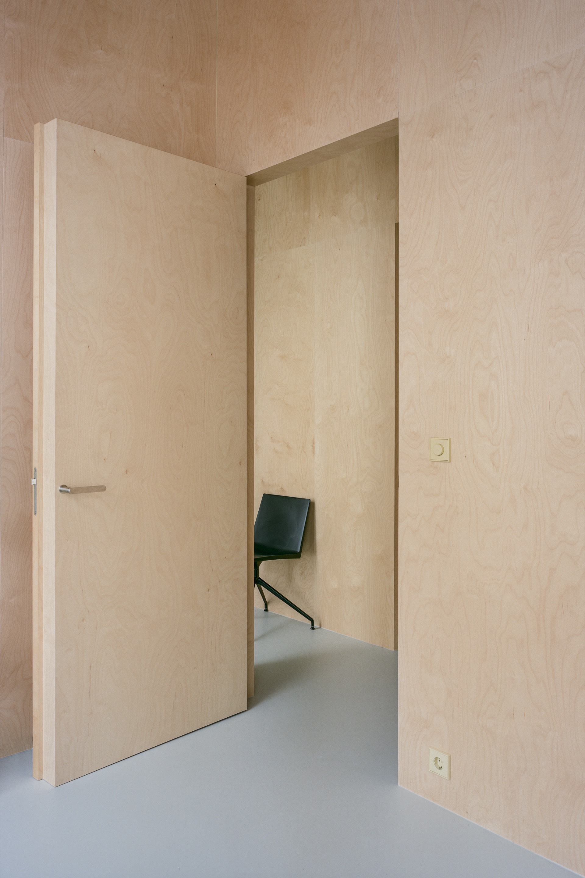 Pale plywood-lined hallway with flush door and glimpse of black chair beyond