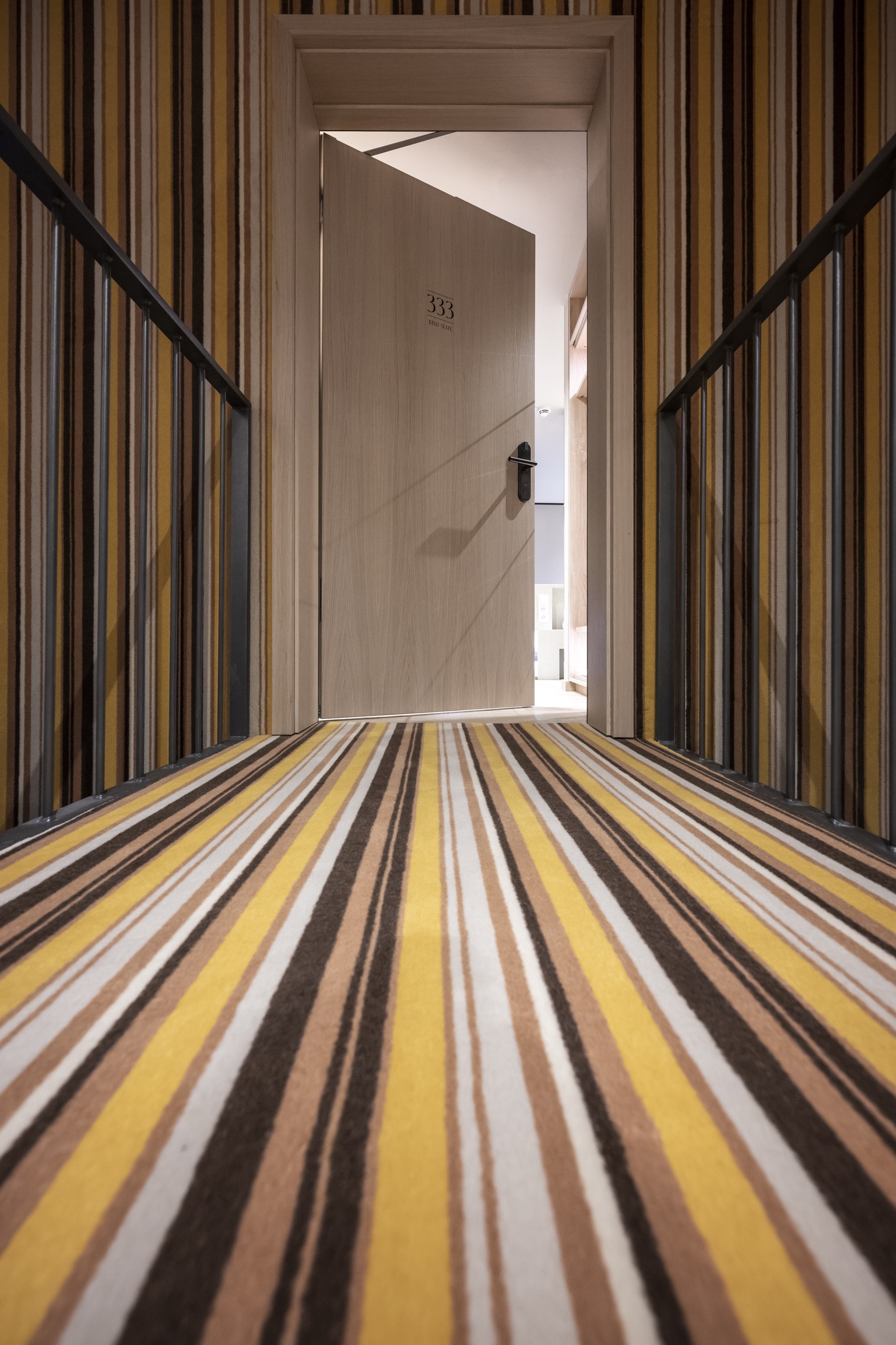 Corridor with striped carpet in yellow and brown tones leading to a numbered door framed by vertical wood slats