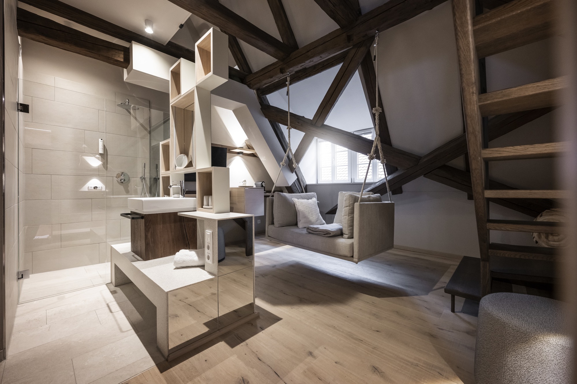 Open bathroom with glass shower enclosure and exposed timber trusses cutting diagonally through the volume