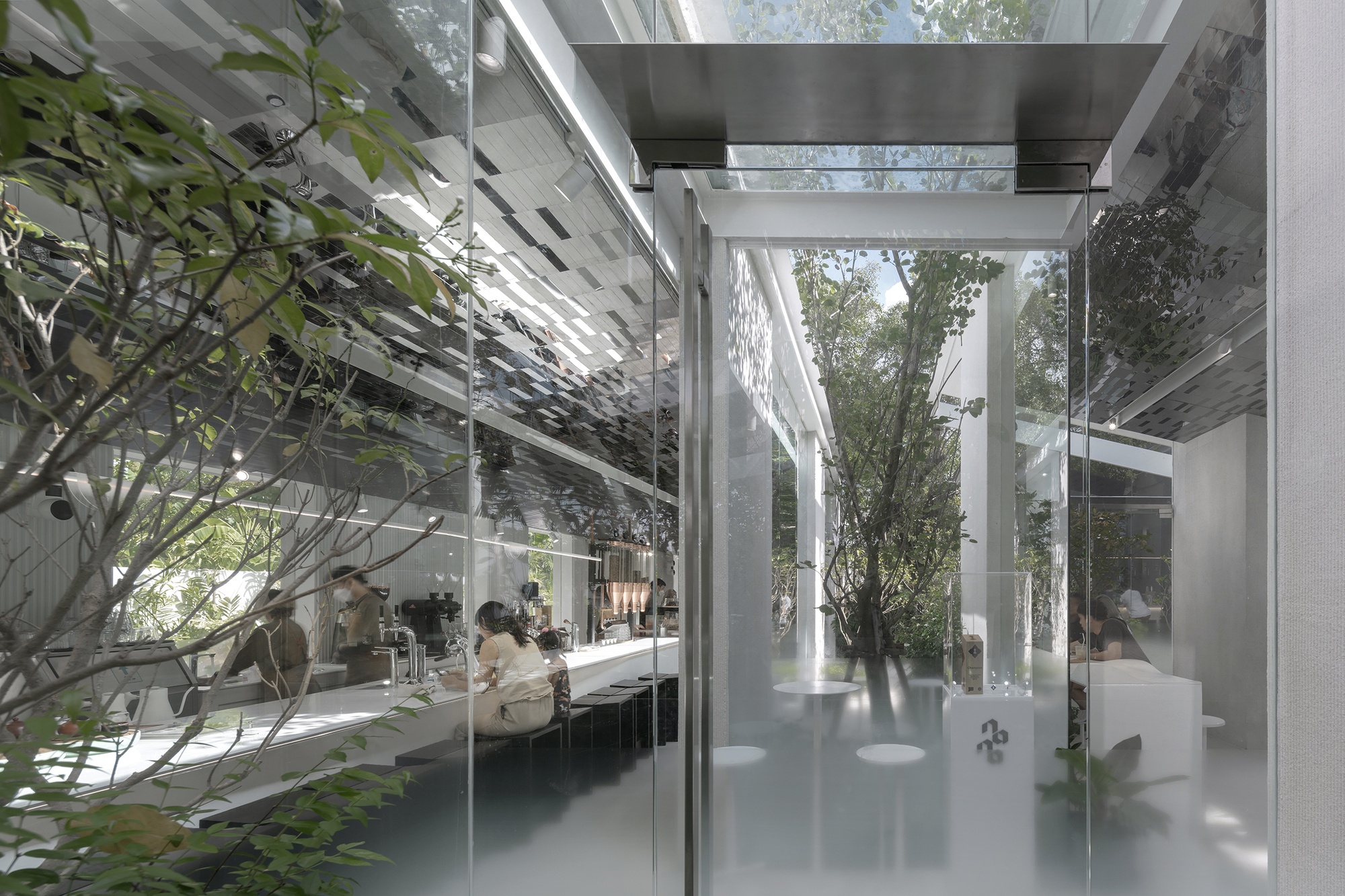 Glass-enclosed interior courtyard with trees and visitors at long white tables beneath a pixelated ceiling