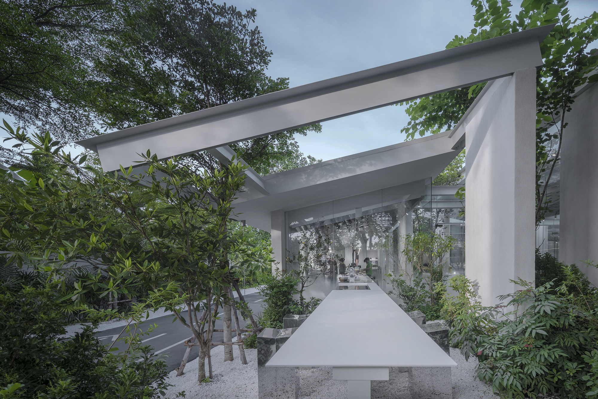 Elevated concrete walkway flanked by planted beds and trees under intersecting white beam structure