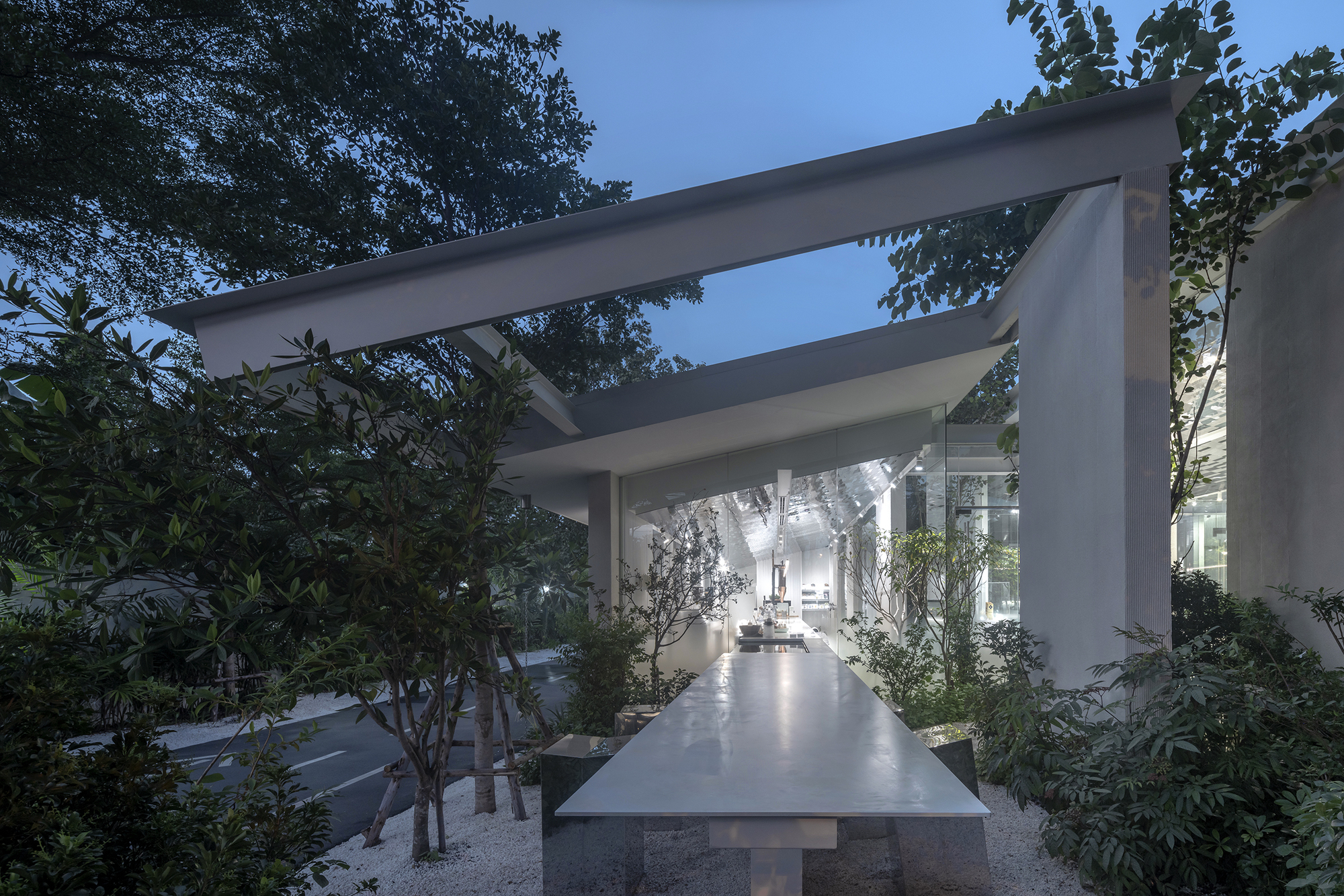 Long concrete table beneath horizontal beam structure surrounded by planted beds at dusk