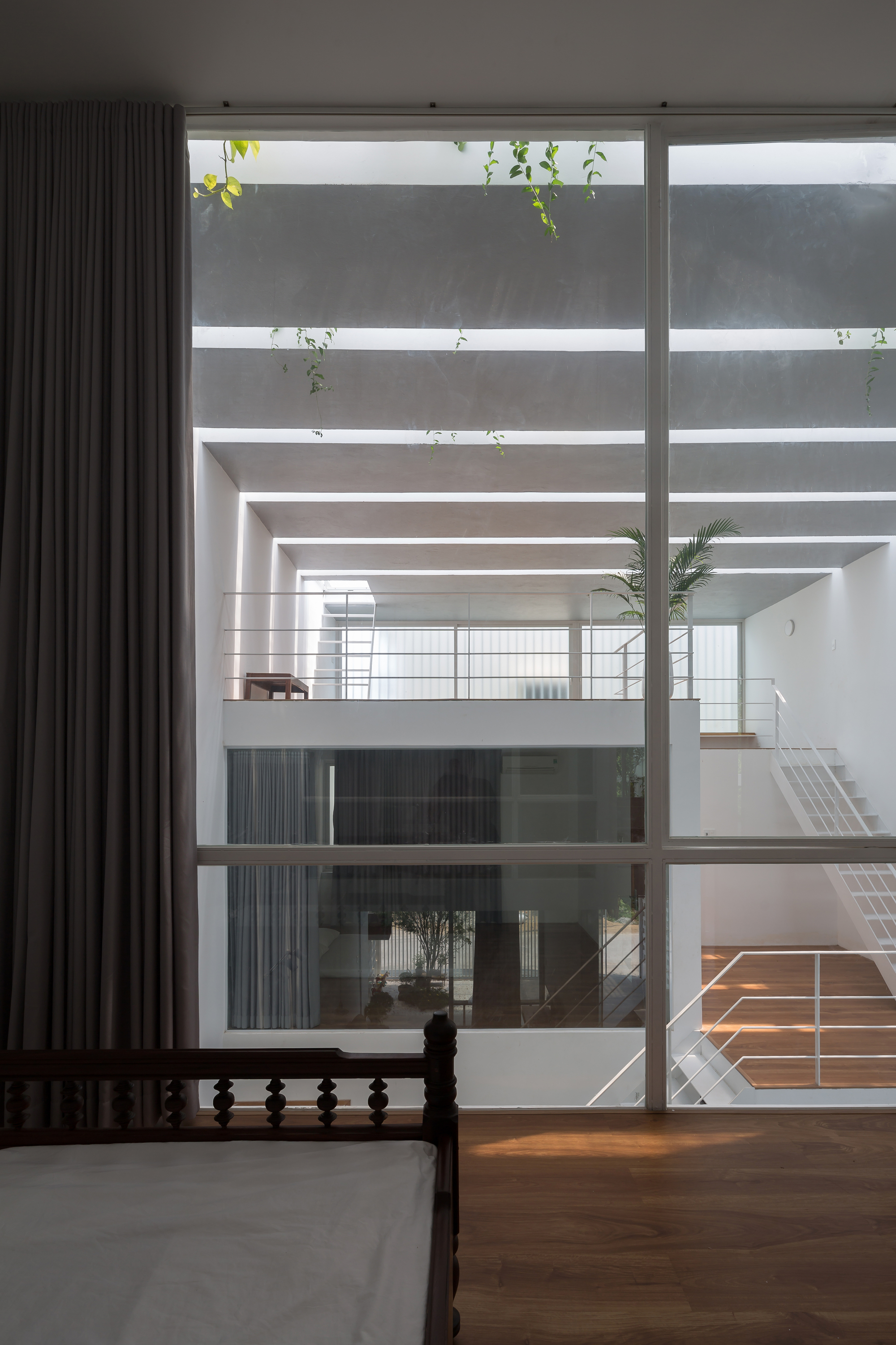 View from bedroom through floor-to-ceiling window into open interior atrium with potted palm and timber floors