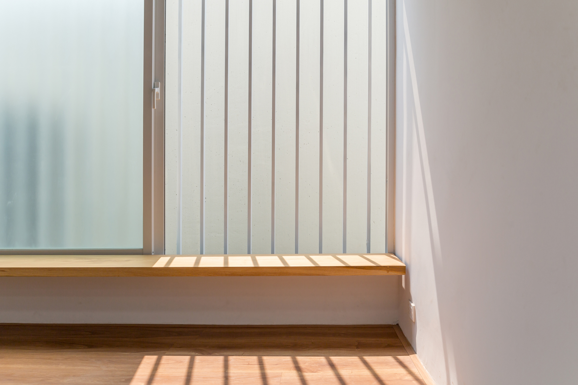 Detail of timber window sill with vertical metal screen casting shadow stripes across wood flooring