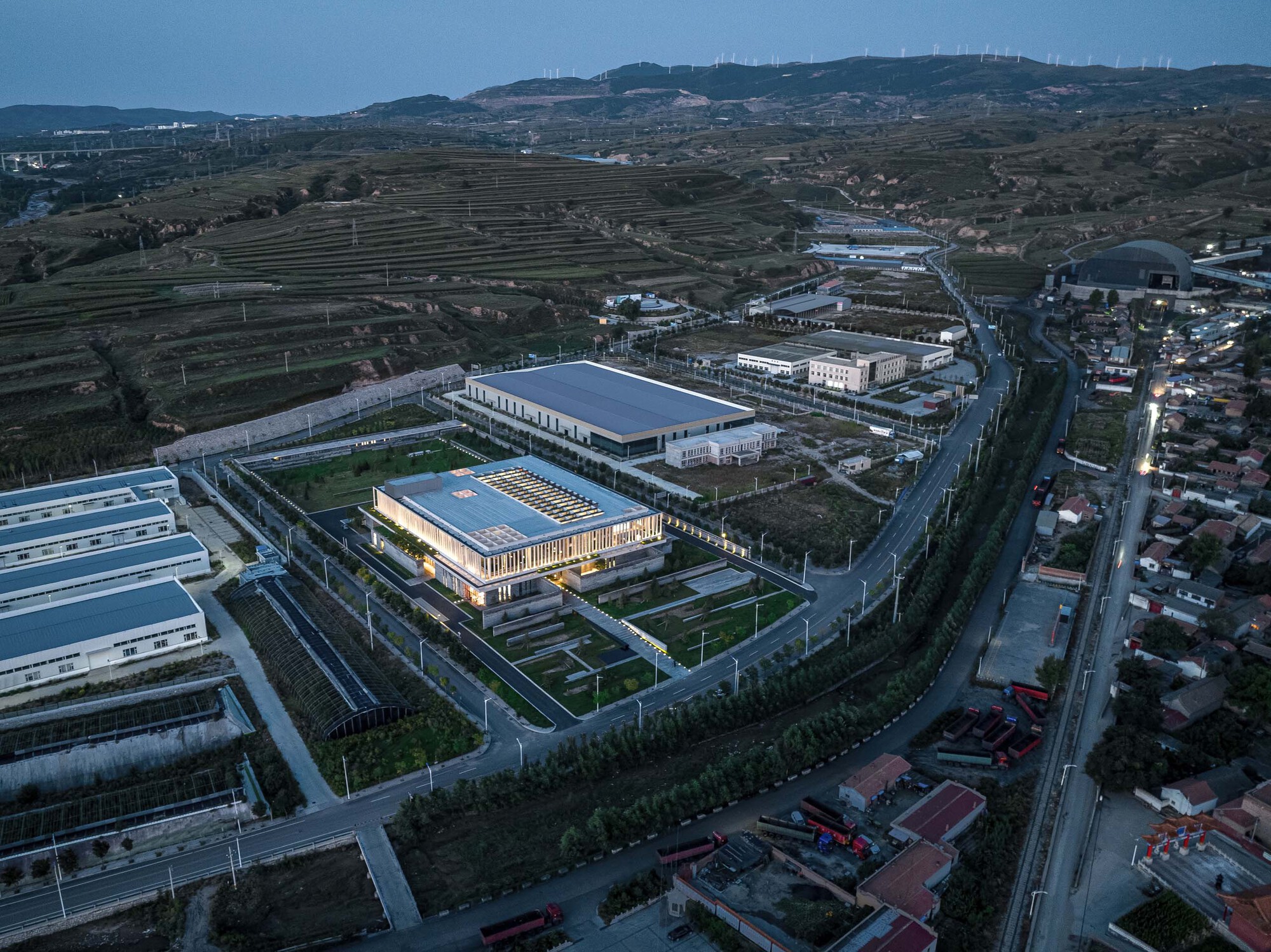 Drone view of the illuminated building surrounded by industrial structures and agricultural terraces at twilight