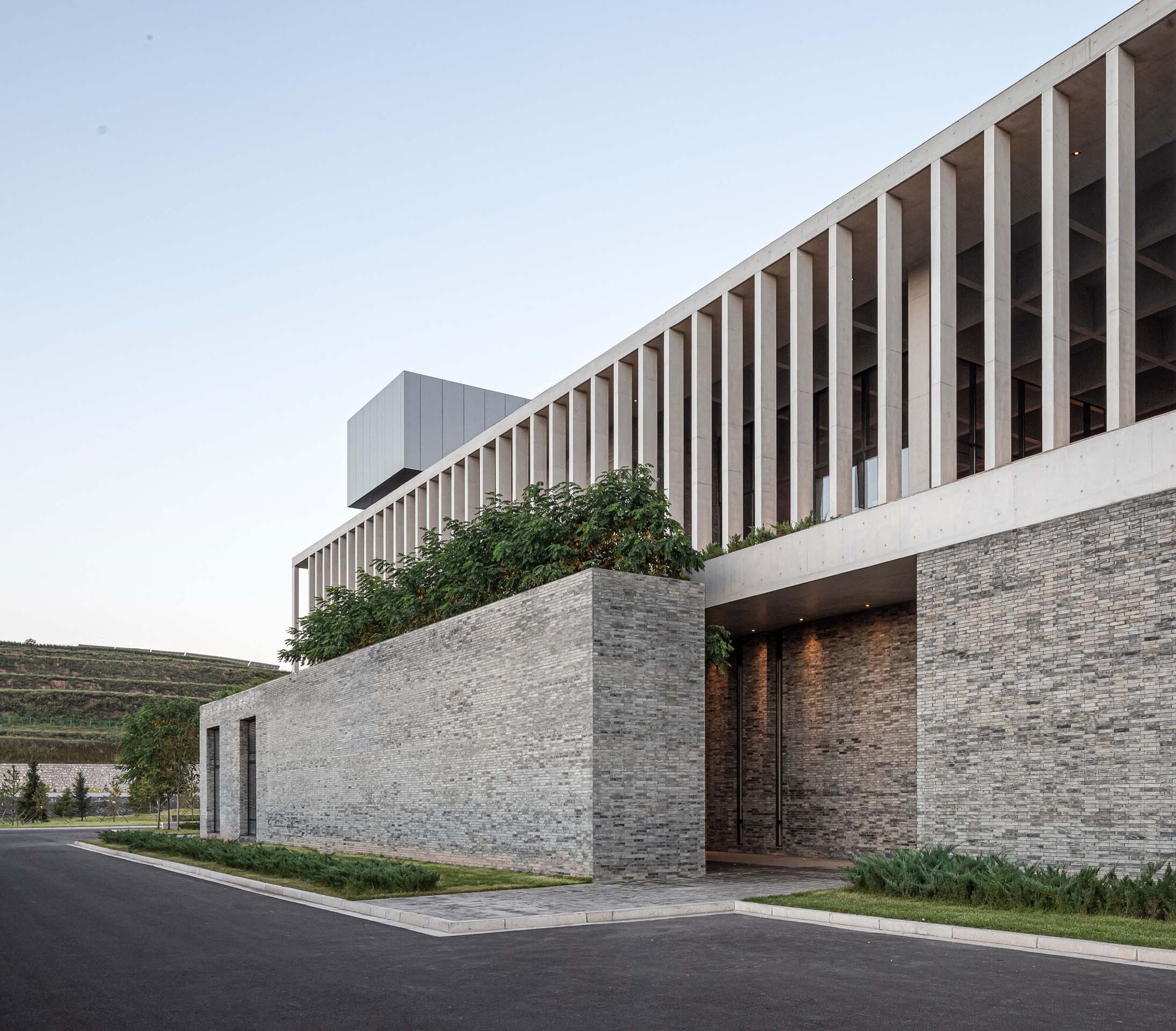 Layered stone entrance wall with recessed doorway beneath the projecting slatted timber volume and climbing greenery