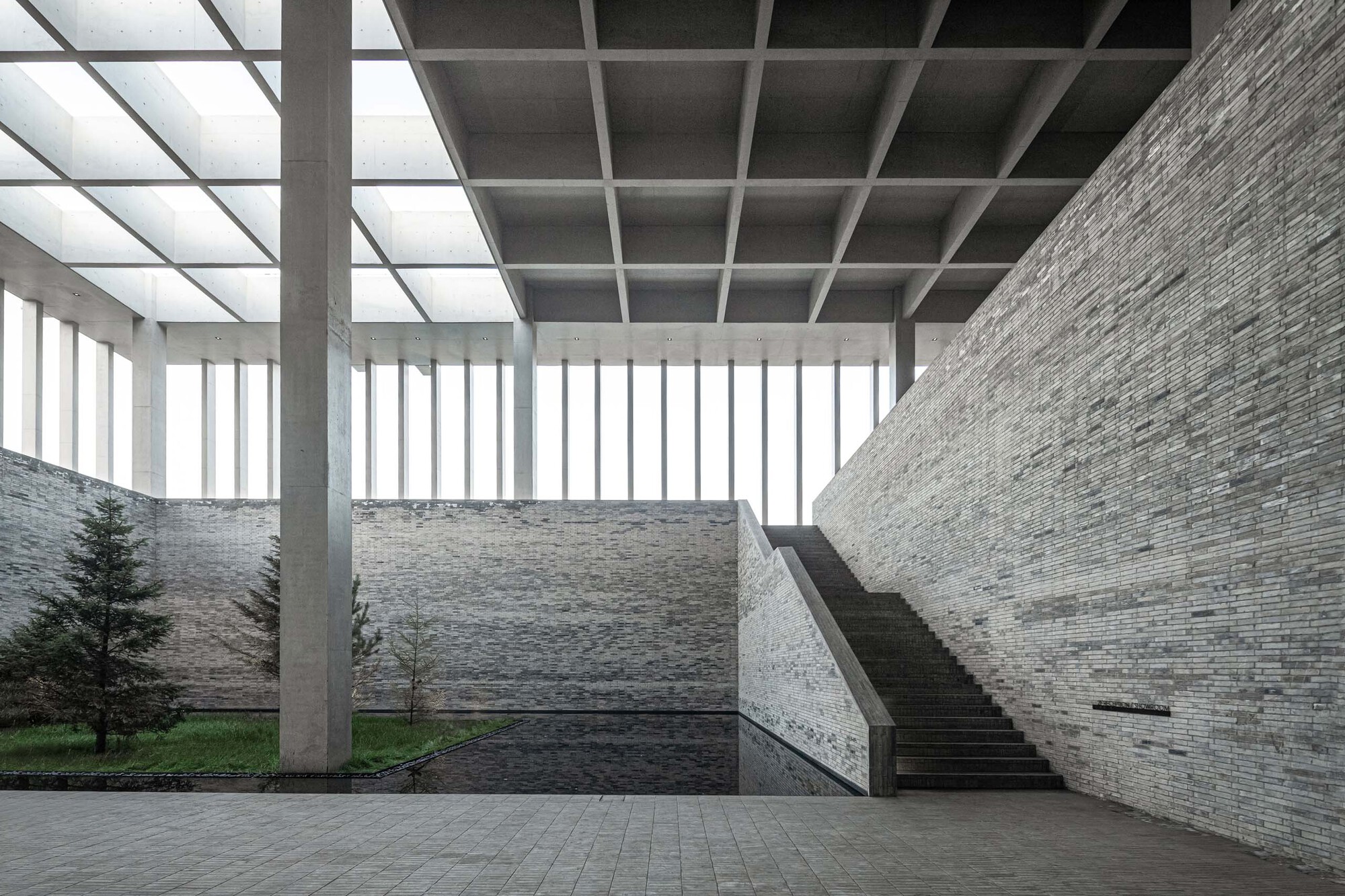 Interior courtyard with stone walls, concrete columns supporting a coffered skylight, and a staircase under diffused light