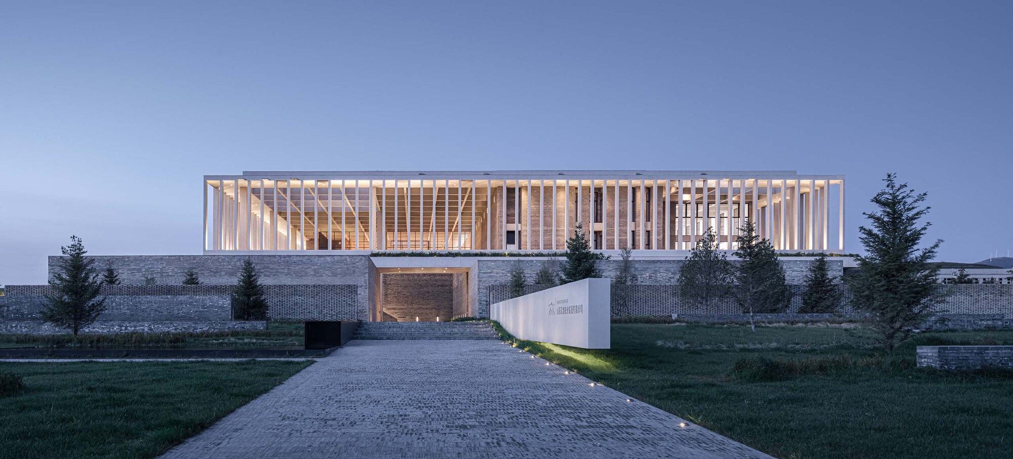 Illuminated vertical louver facade atop a stone plinth with a gravel entry path at dusk