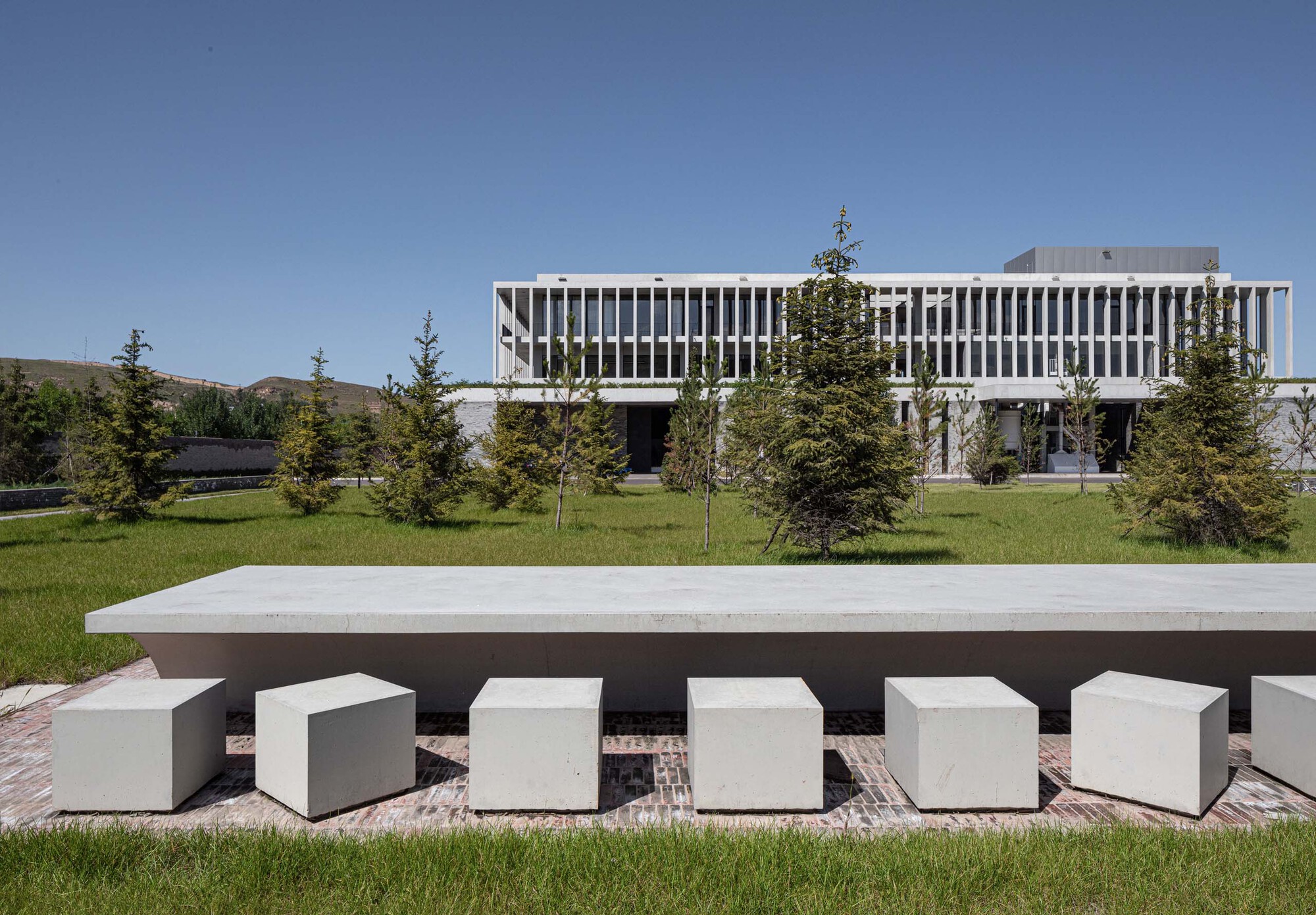Outdoor seating area with concrete table and cube stools on a lawn before the vertical-louvered facade