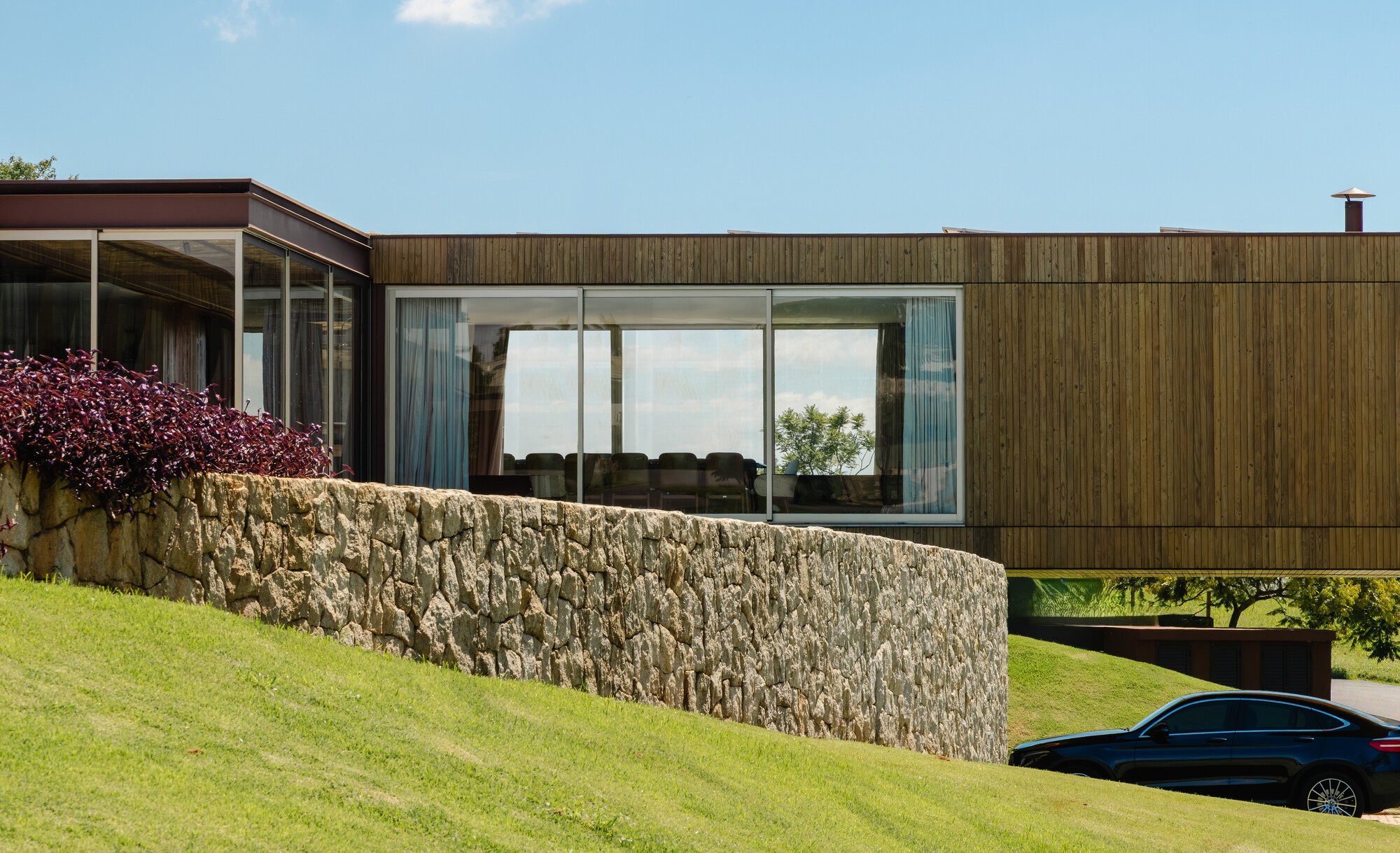 Cantilevered timber-clad volume above fieldstone retaining wall with planted bed and parked car below