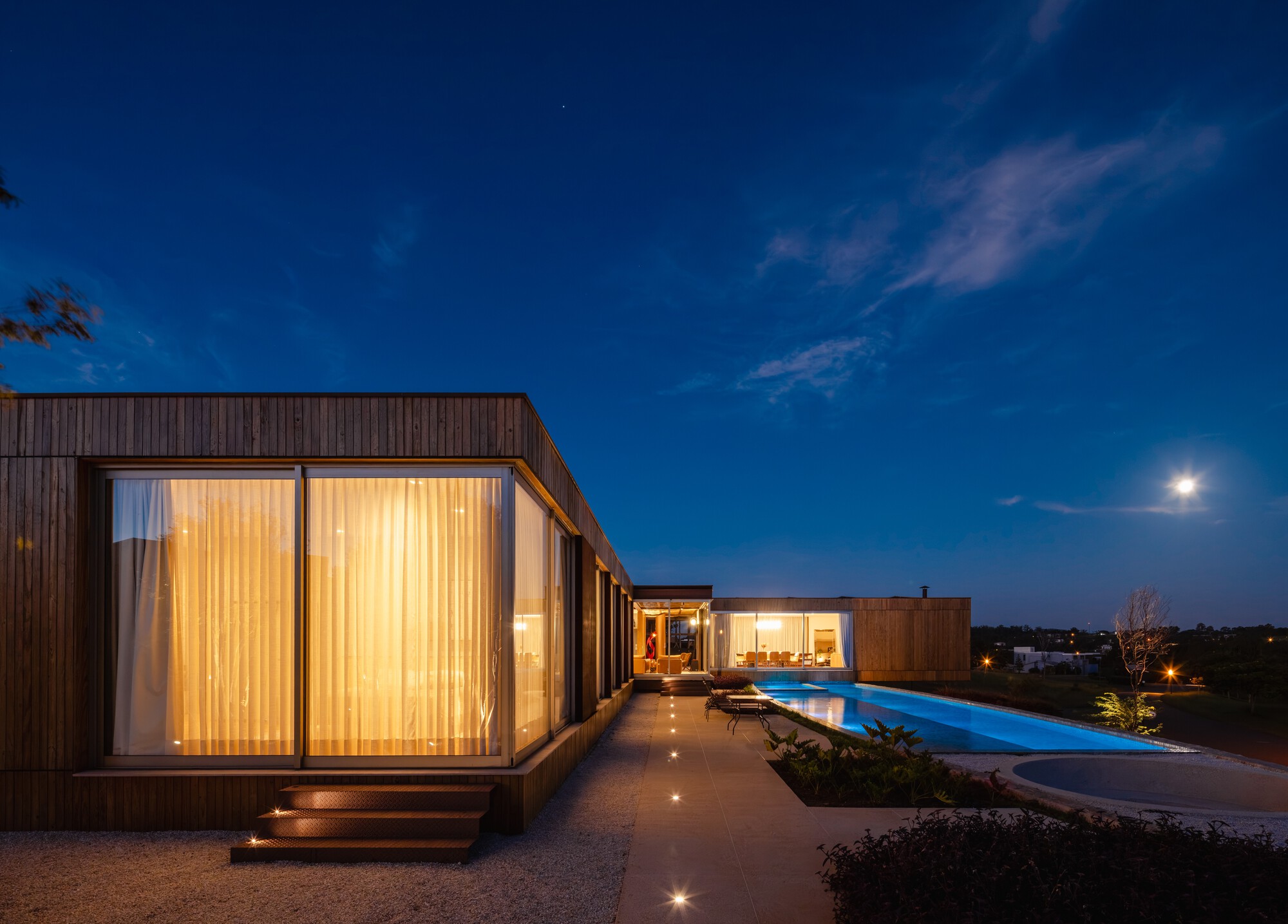 Glazed bedroom volume with curtains beside a pool and planted beds at dusk