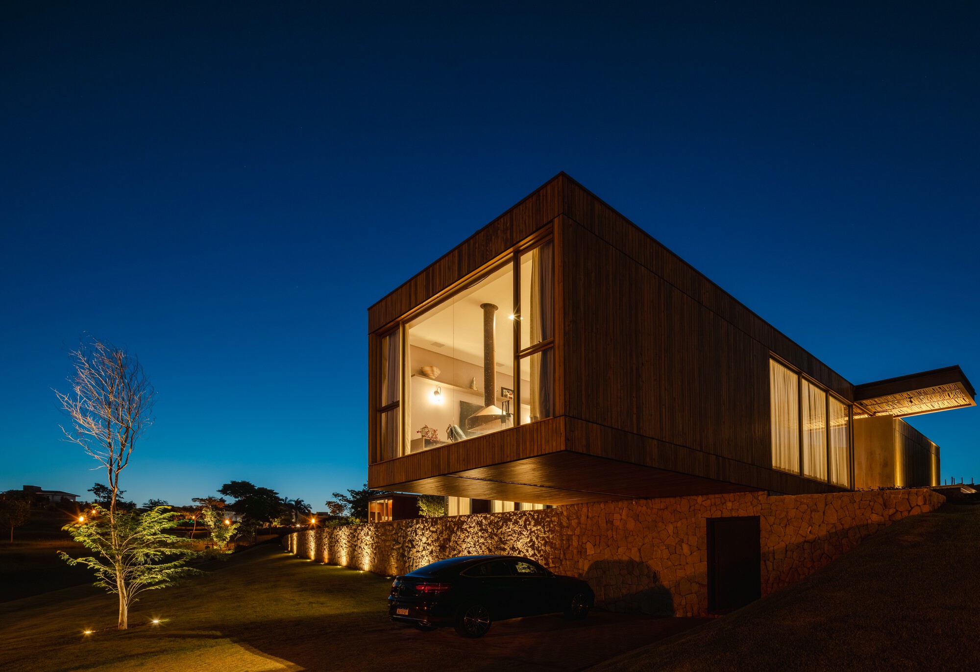 Corner-glazed timber volume cantilevered above a stone retaining wall at night