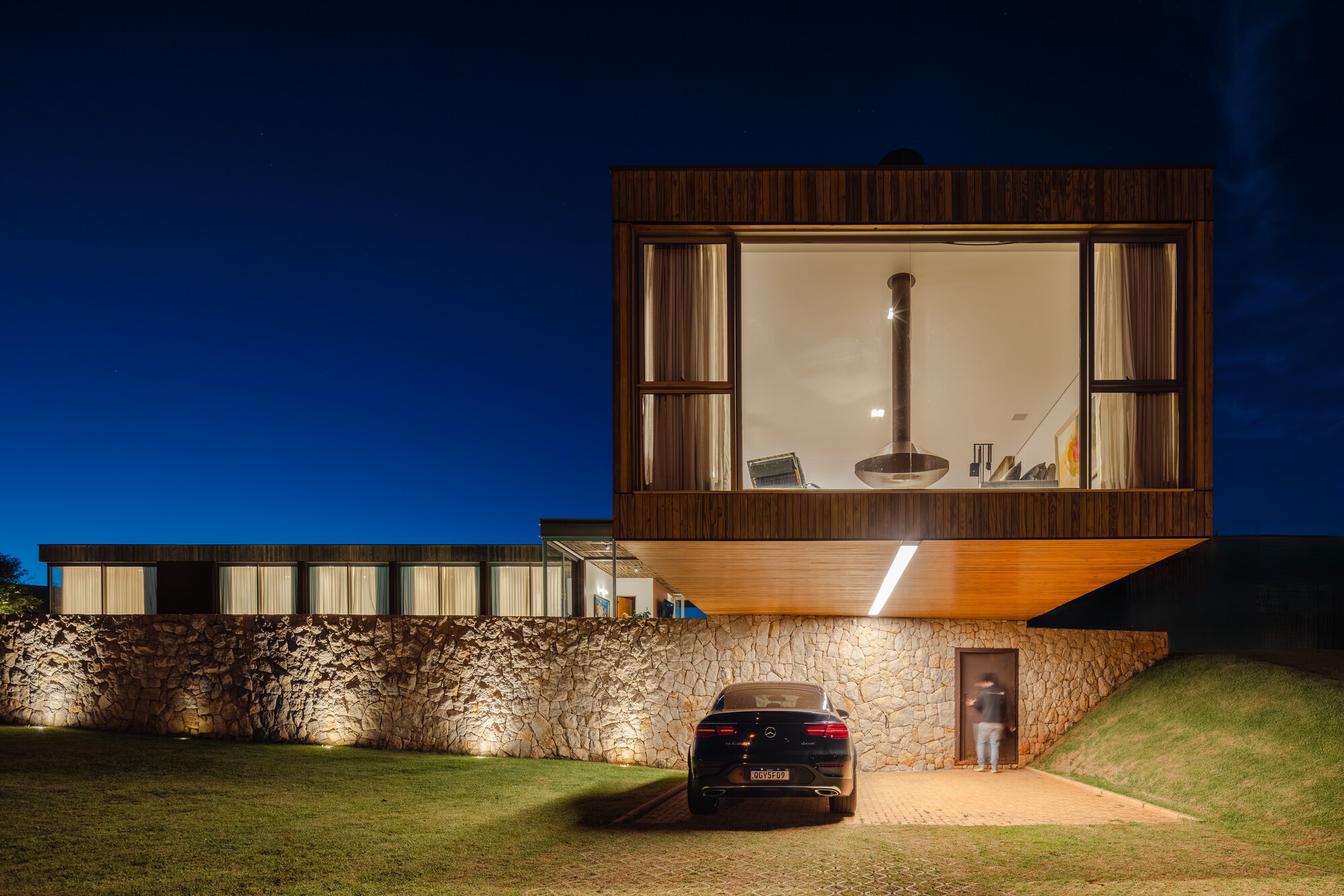 Cantilevered timber box above a stone base with a carport entry at twilight