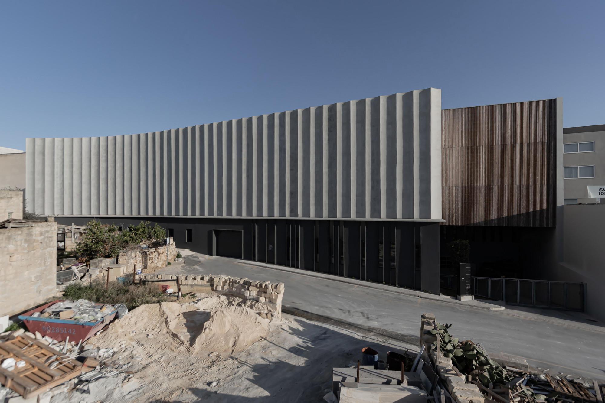 Facade with white vertical louvres and timber-clad volume rising above a paved forecourt under clear sky