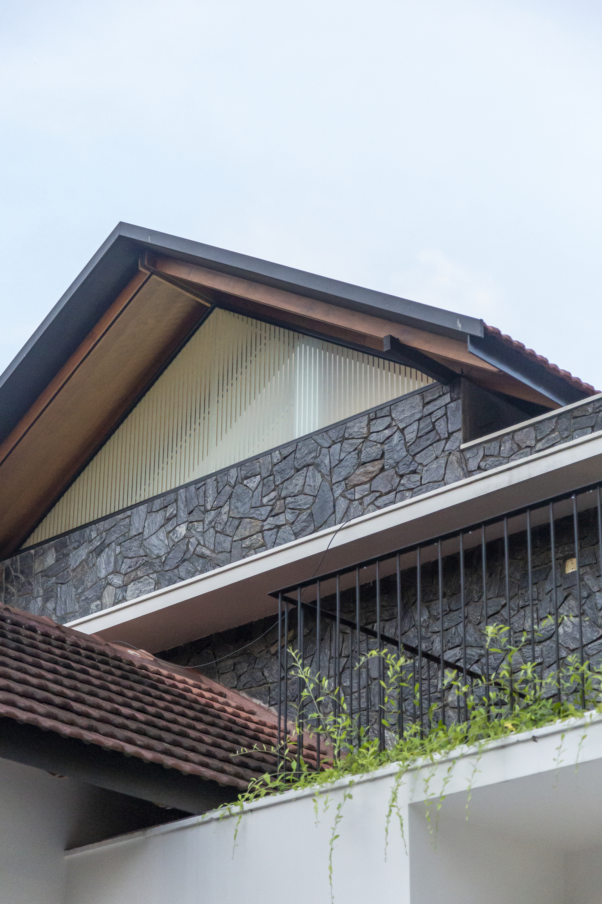Upper level facade with corrugated metal gable panel, stone balcony parapet, and planters below a cloudy sky