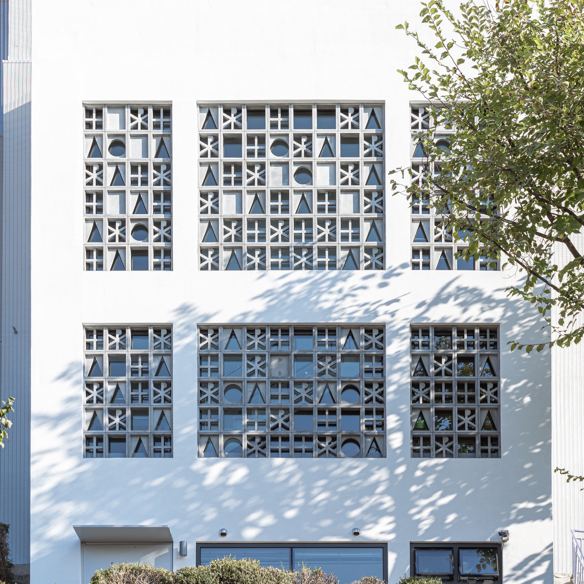 White stucco facade with geometric patterned window screens casting dappled tree shadows in daylight