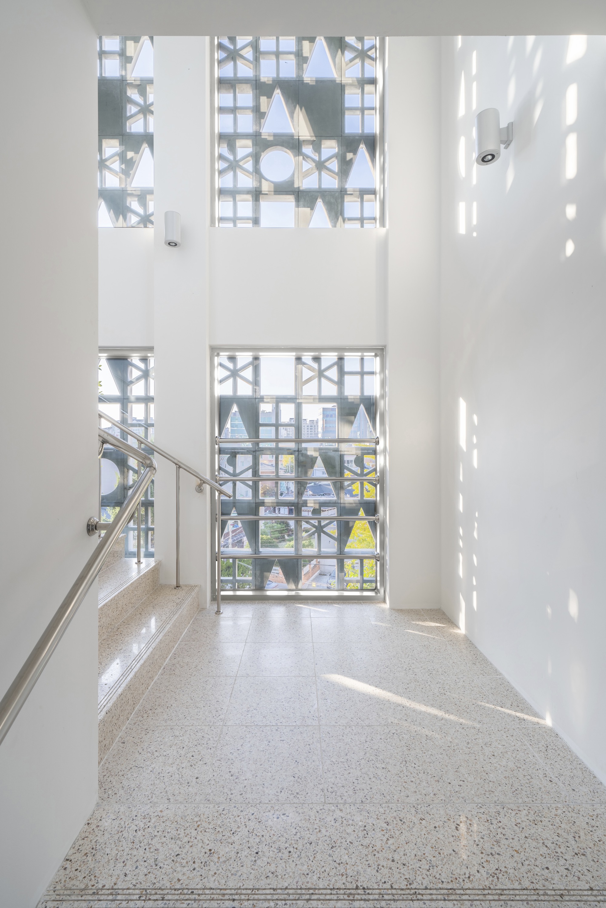 Double-height stairwell with terrazzo flooring and geometric window screens filtering bright daylight patterns
