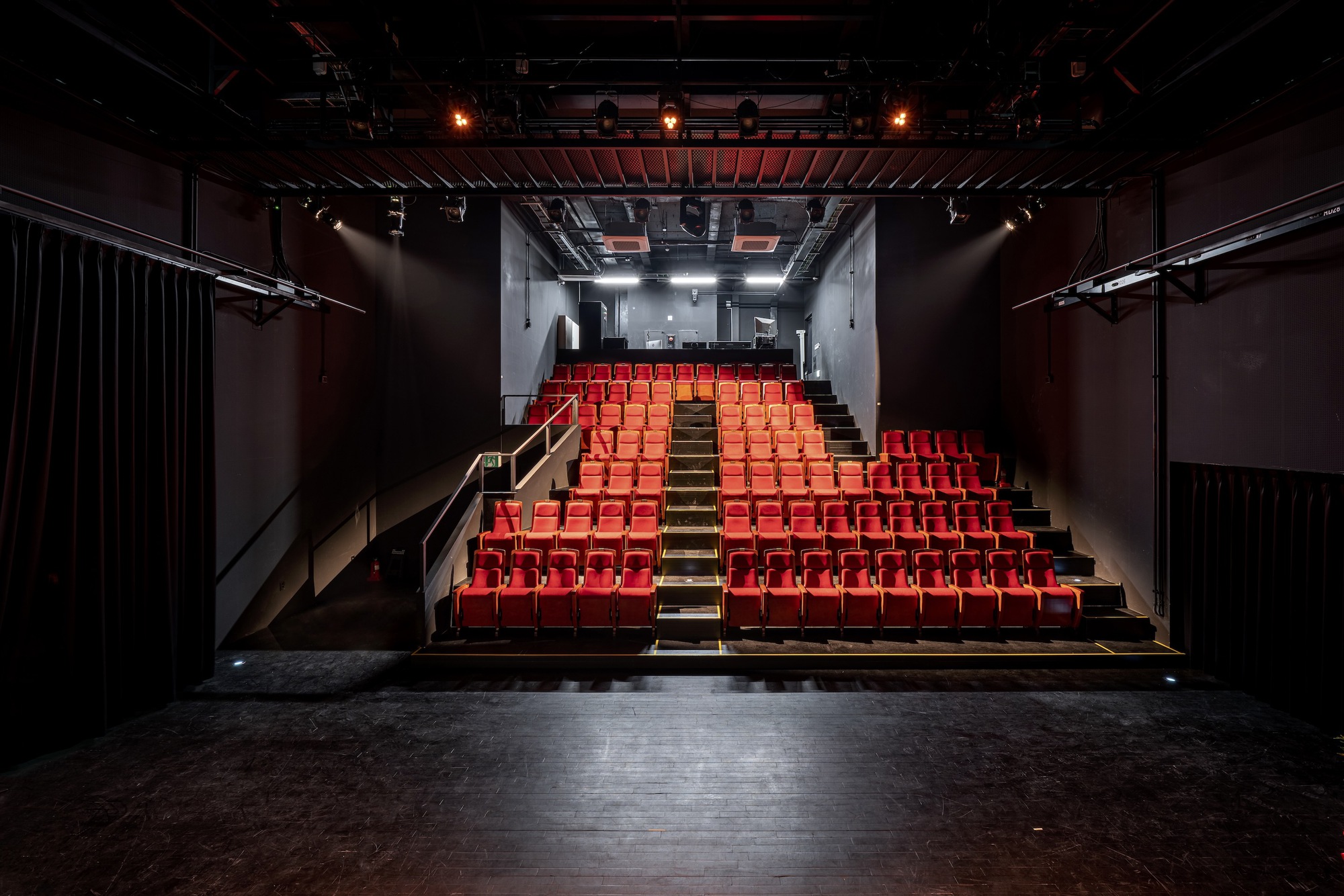 View from the stage toward the tiered red seating under exposed black ceiling trusses and lighting rigs