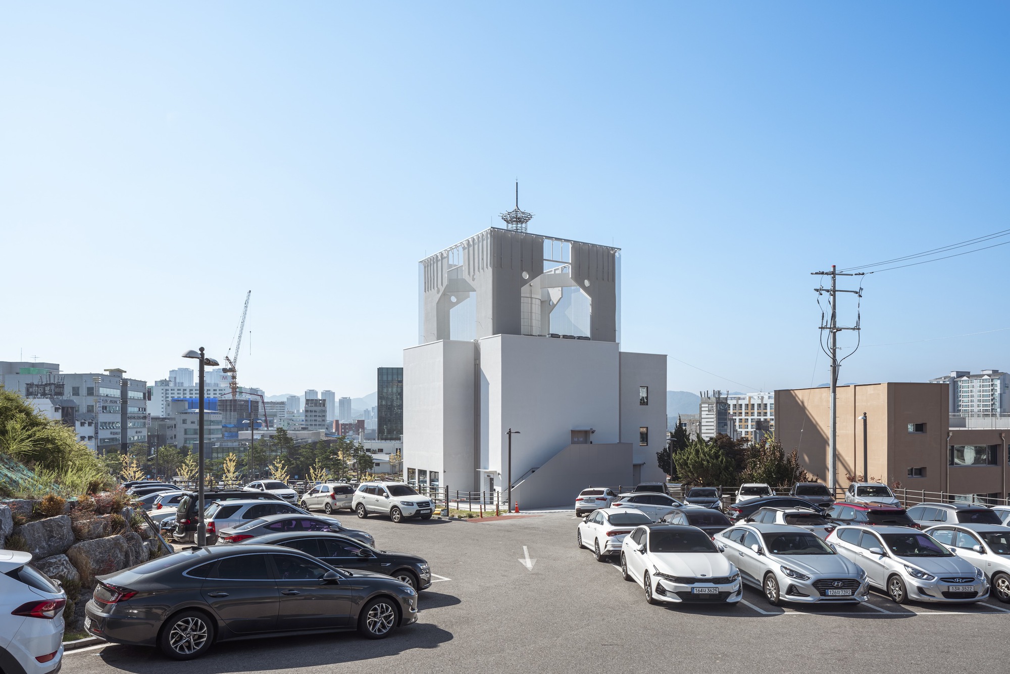 White tower with arched openings rising above a parking lot filled with cars under clear blue sky