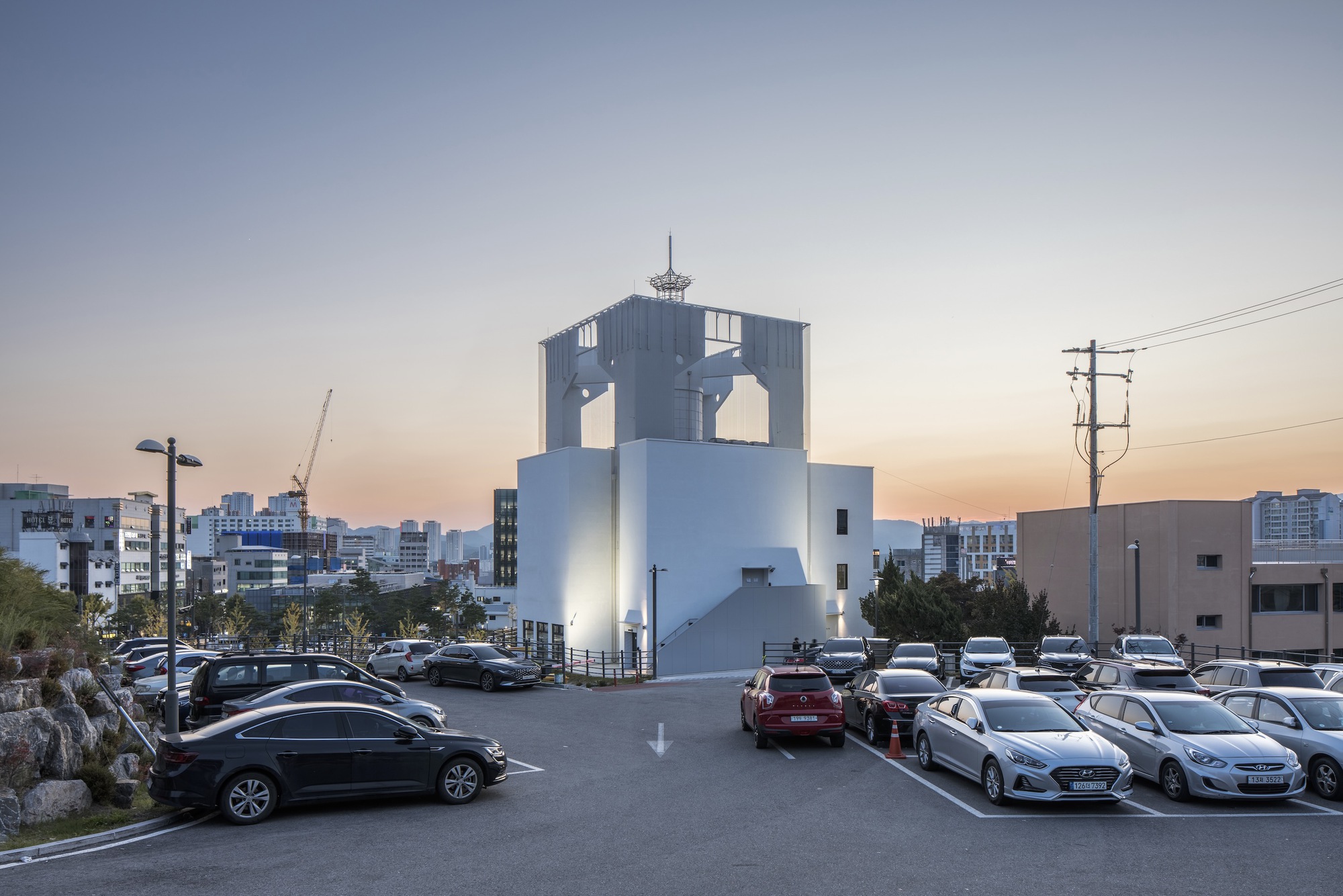 White tower with arched openings illuminated at dusk beside a parking lot and utility poles