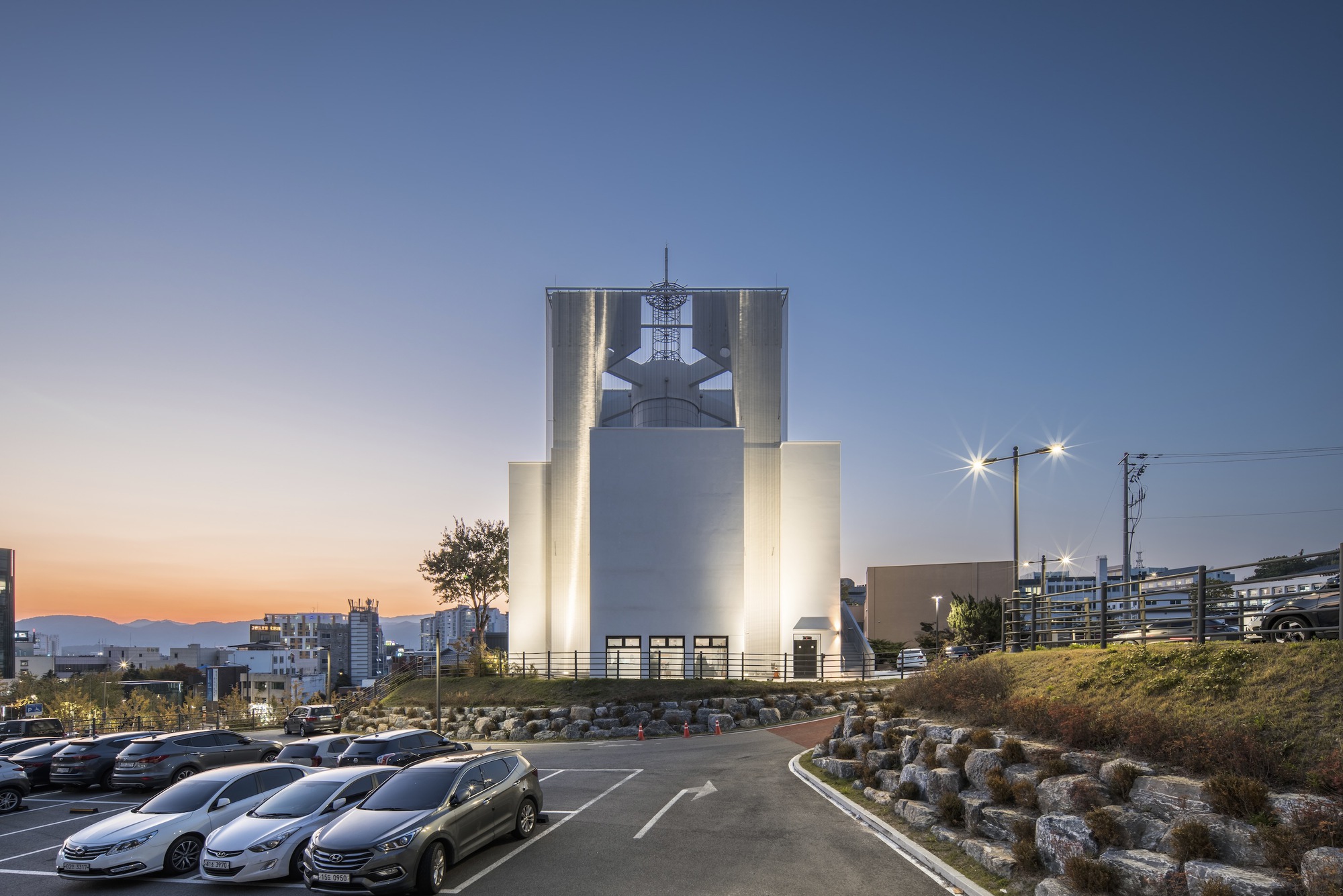 Illuminated cylindrical tower housing a bell in front of a parking area at sunset