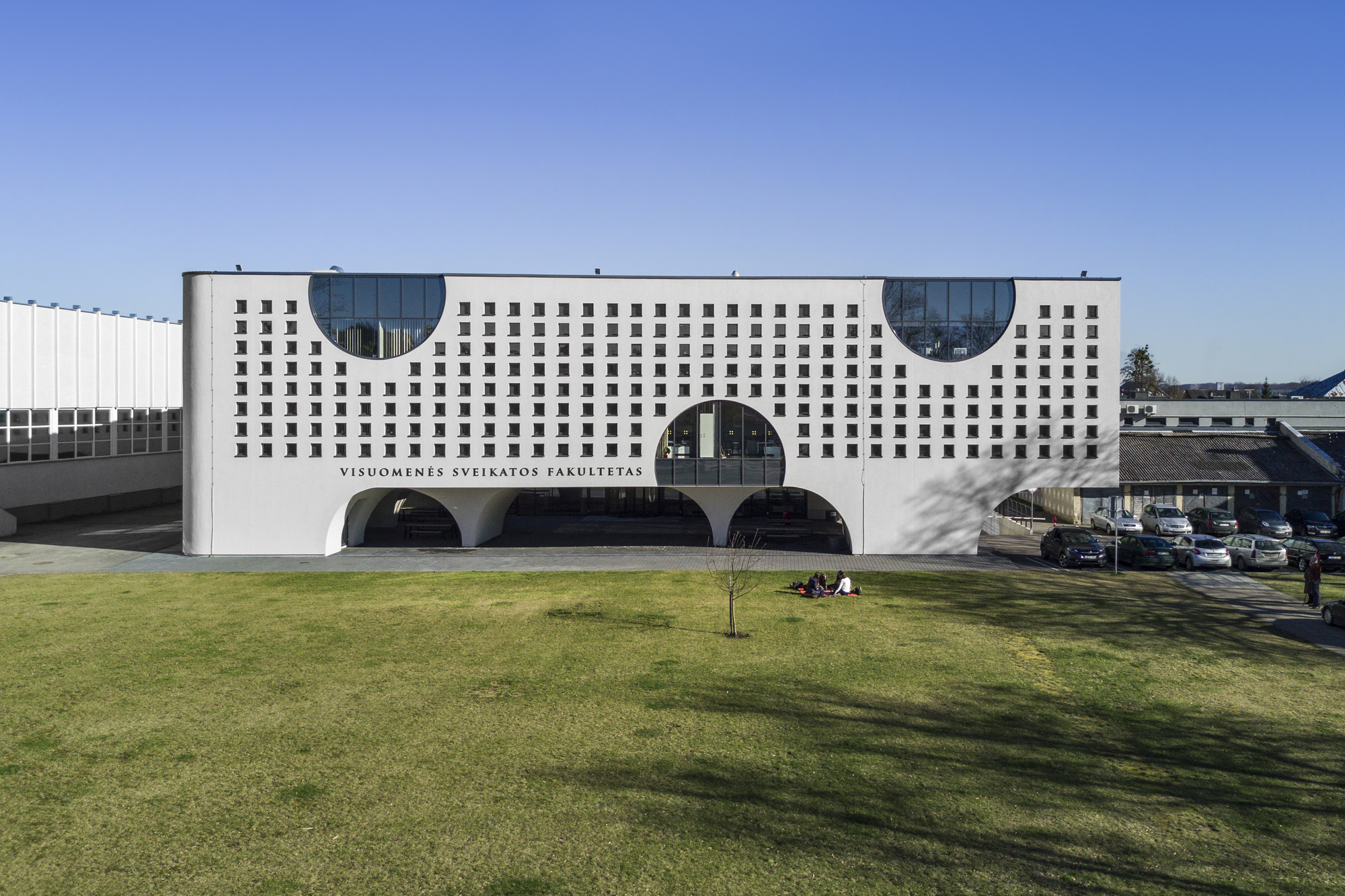 White perforated facade with circular glazed cutouts and arched openings at ground level across a green lawn