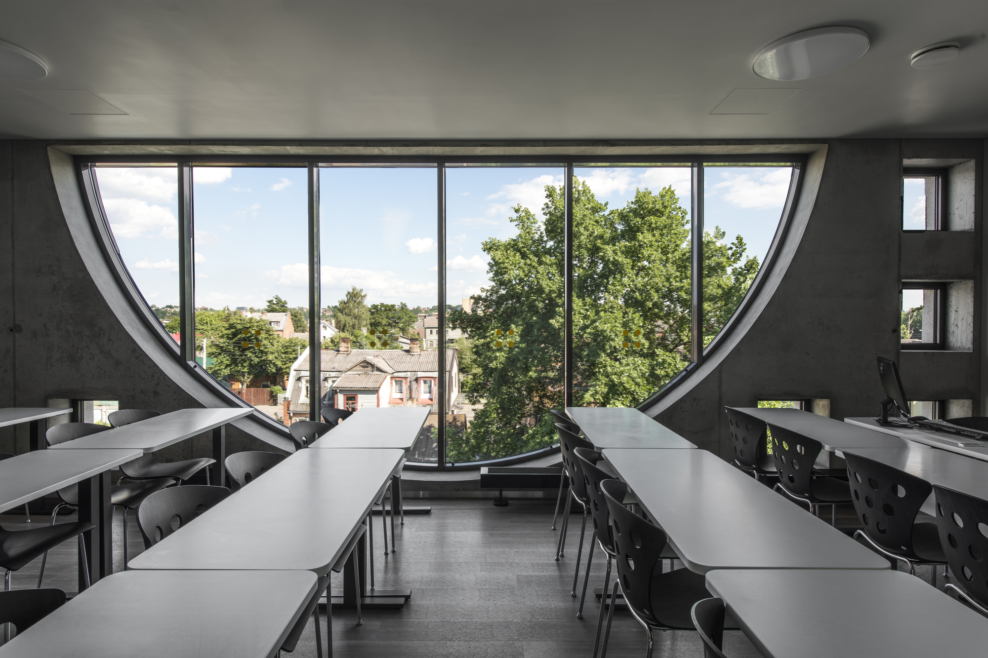 Interior classroom with rows of tables facing a semicircular window overlooking leafy trees and rooftops