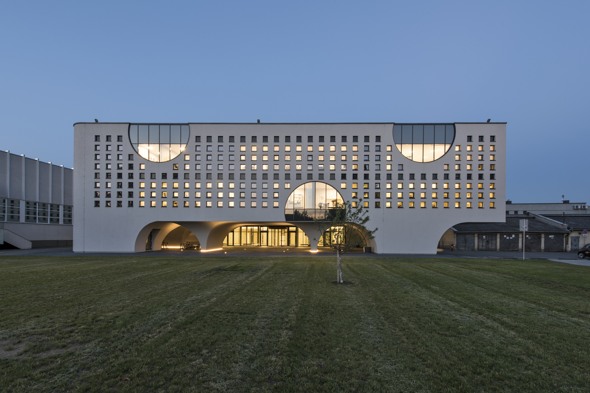 Front elevation showing the gridded white facade with illuminated arched entrance portals at dusk