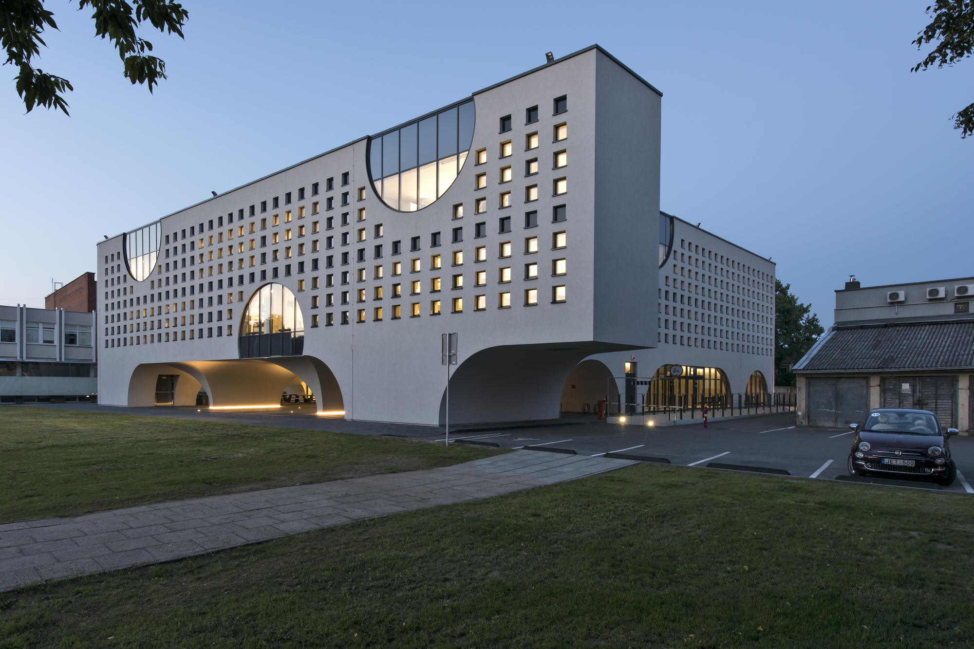 Street view of the white perforated facade with lit arched entrances beneath trees at twilight
