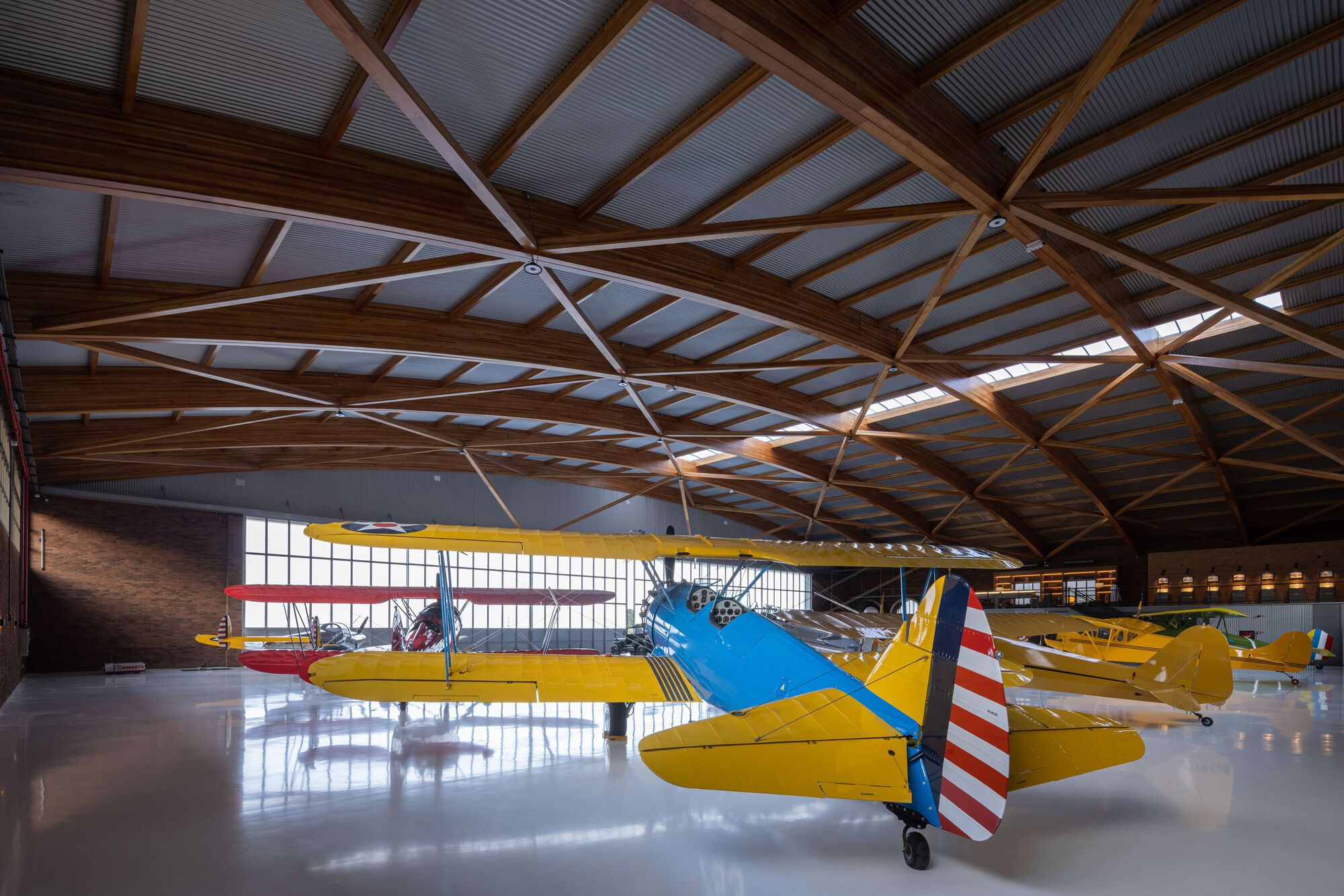 Interior view of timber and metal truss roof structure over a hangar floor with yellow biplanes