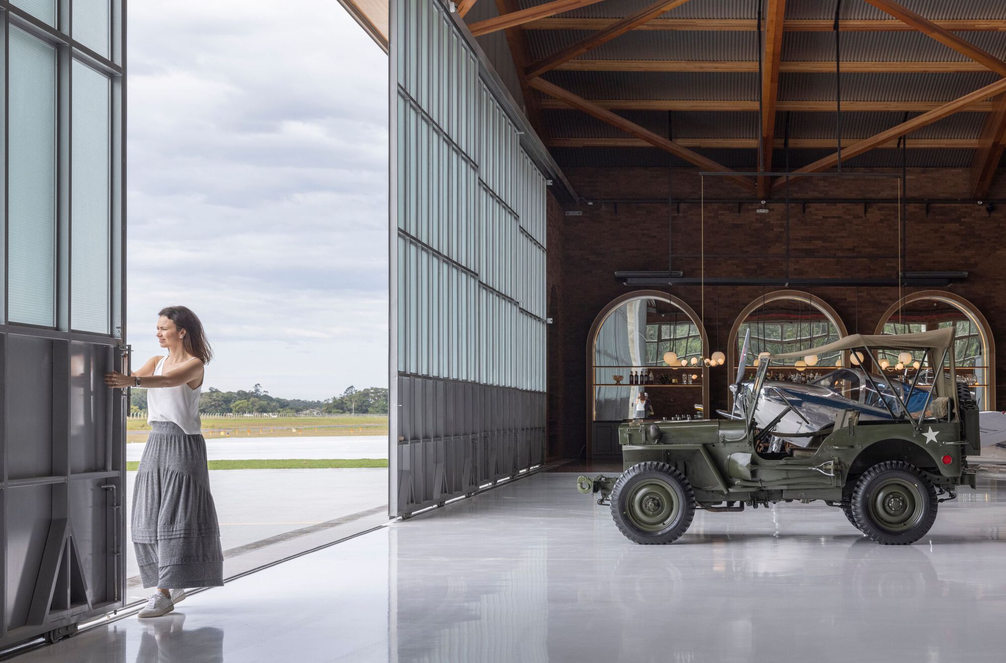 Hangar interior with corrugated metal walls, timber roof trusses, and a vintage military vehicle on polished concrete