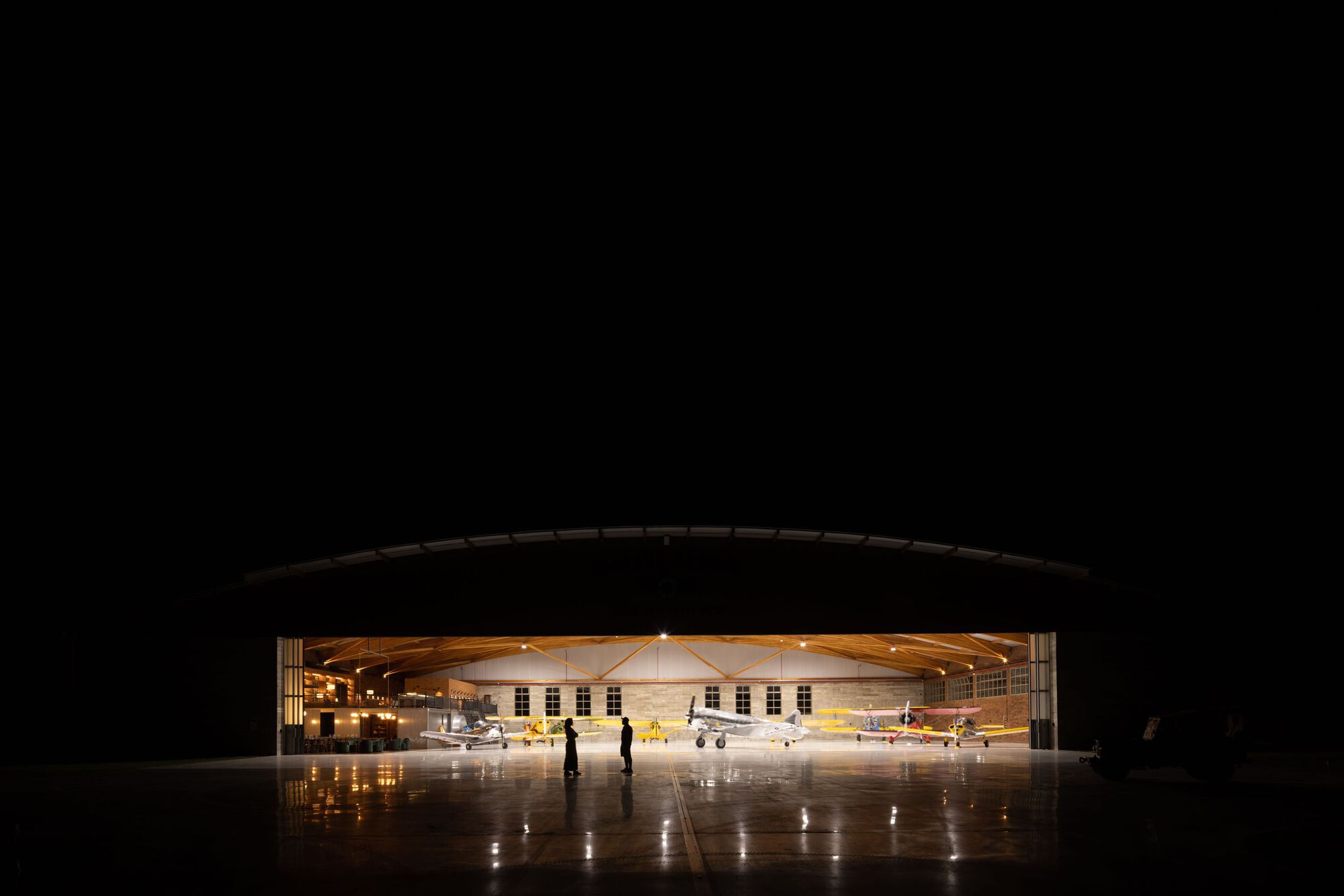Aircraft hangar interior with exposed timber trusses and parked planes viewed through the open door at night