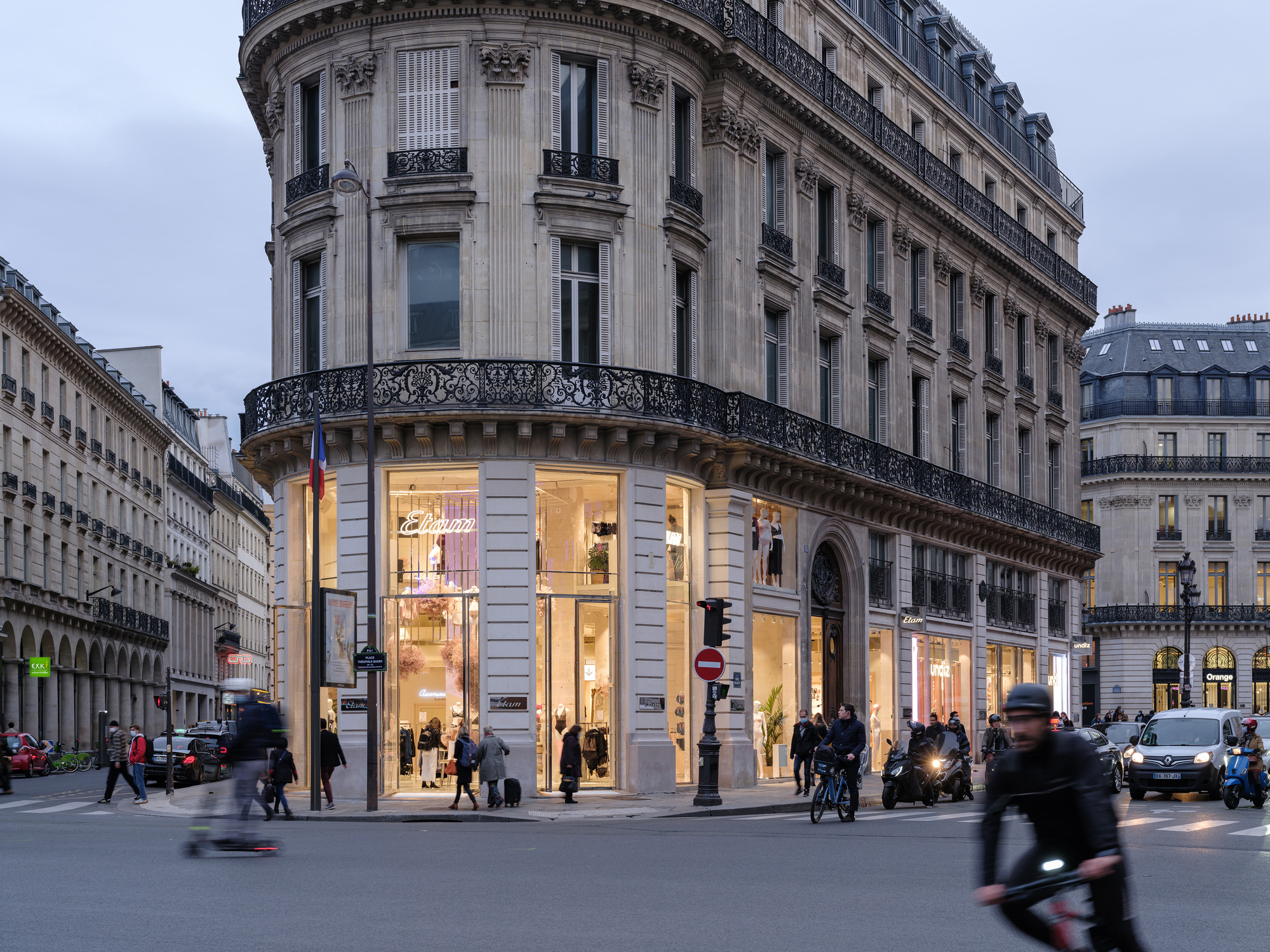 Corner building with rusticated stone base and tall storefront windows glowing at dusk with pedestrians crossing