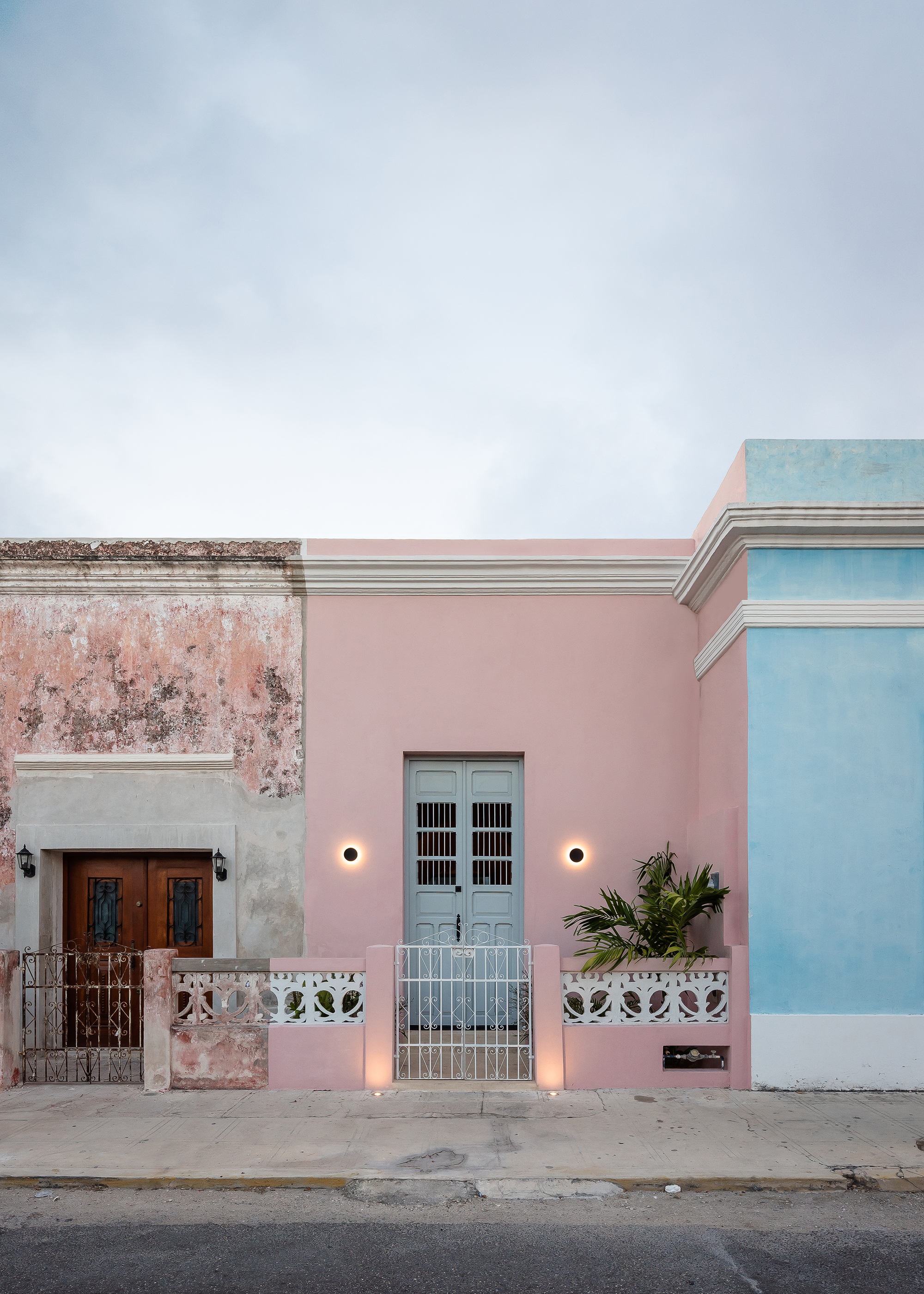 Street facade with pink stucco wall, pale blue door, sconce lighting and weathered neighboring building at dusk
