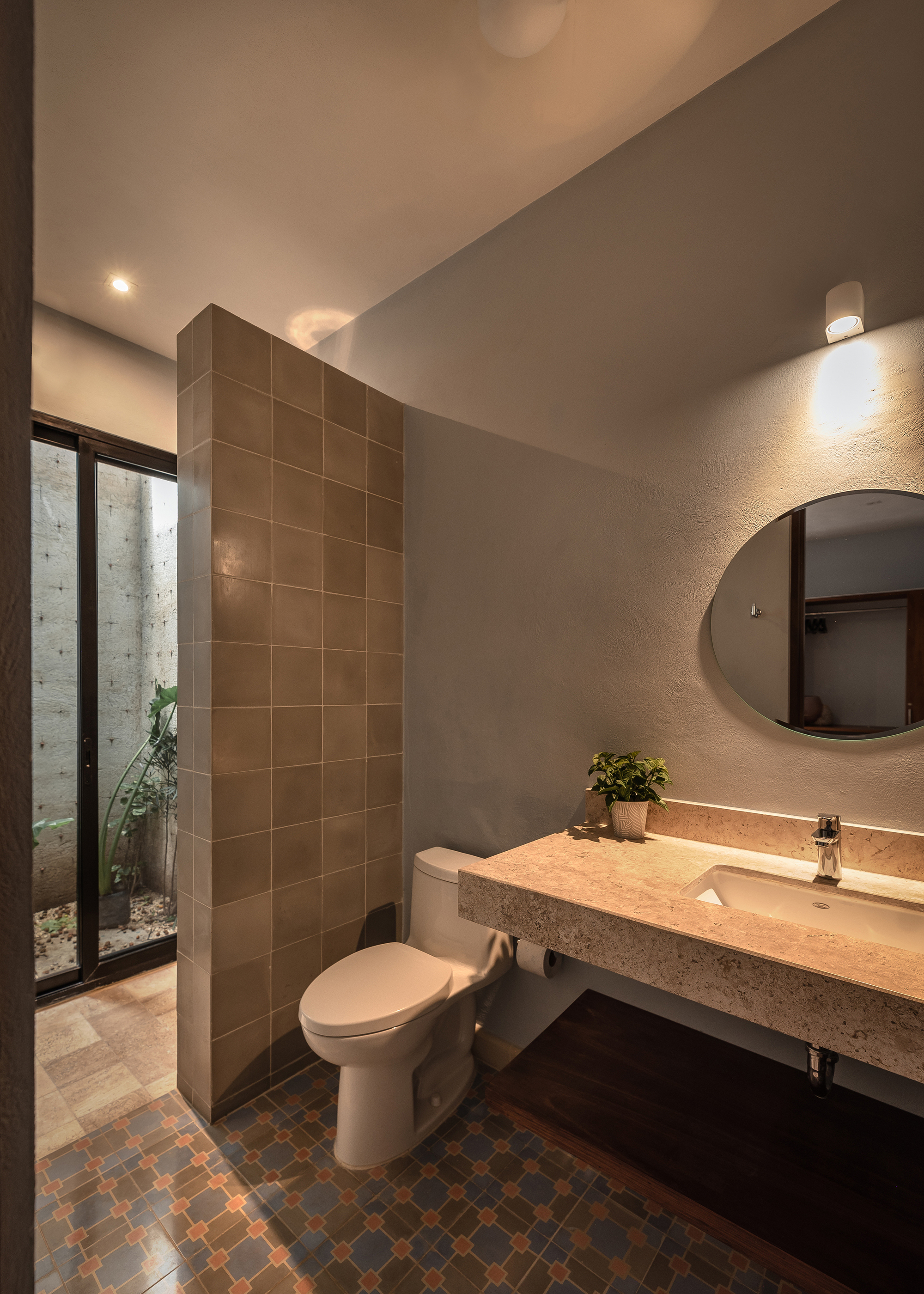 Bathroom interior with brown tile partition wall, terrazzo vanity, and view to planted courtyard through glazed door