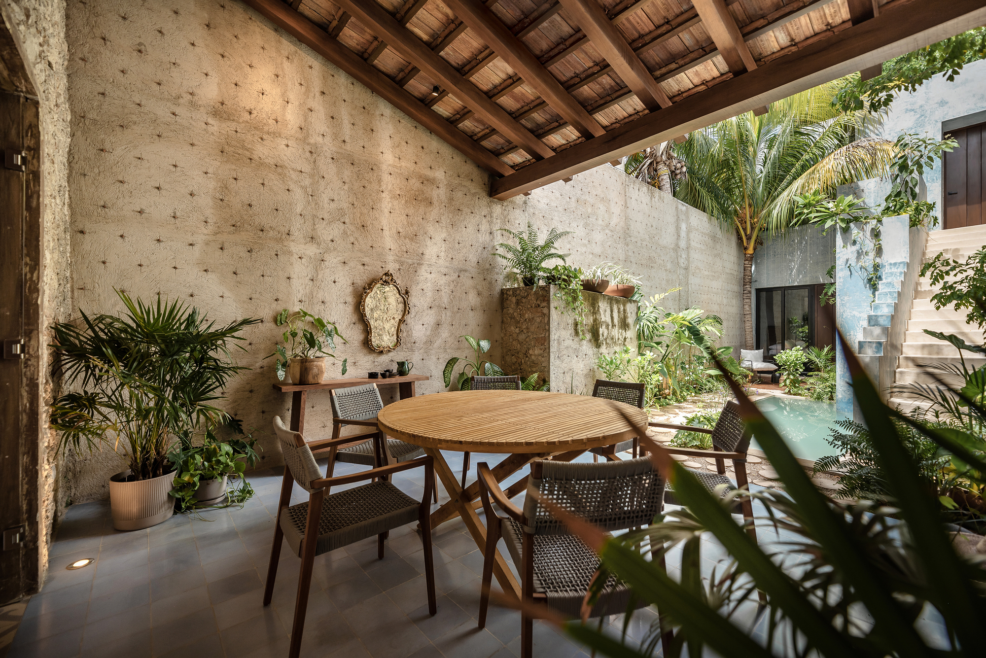 Covered outdoor dining terrace with timber beam ceiling and concrete walls opening to a planted garden