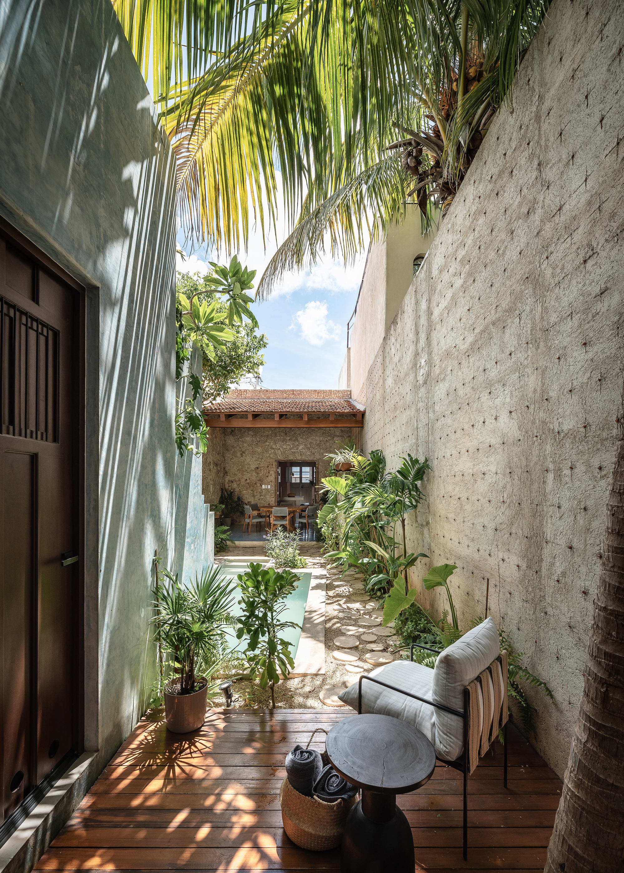 Narrow courtyard with board-formed concrete walls, timber deck seating area, and potted palms under dappled sunlight