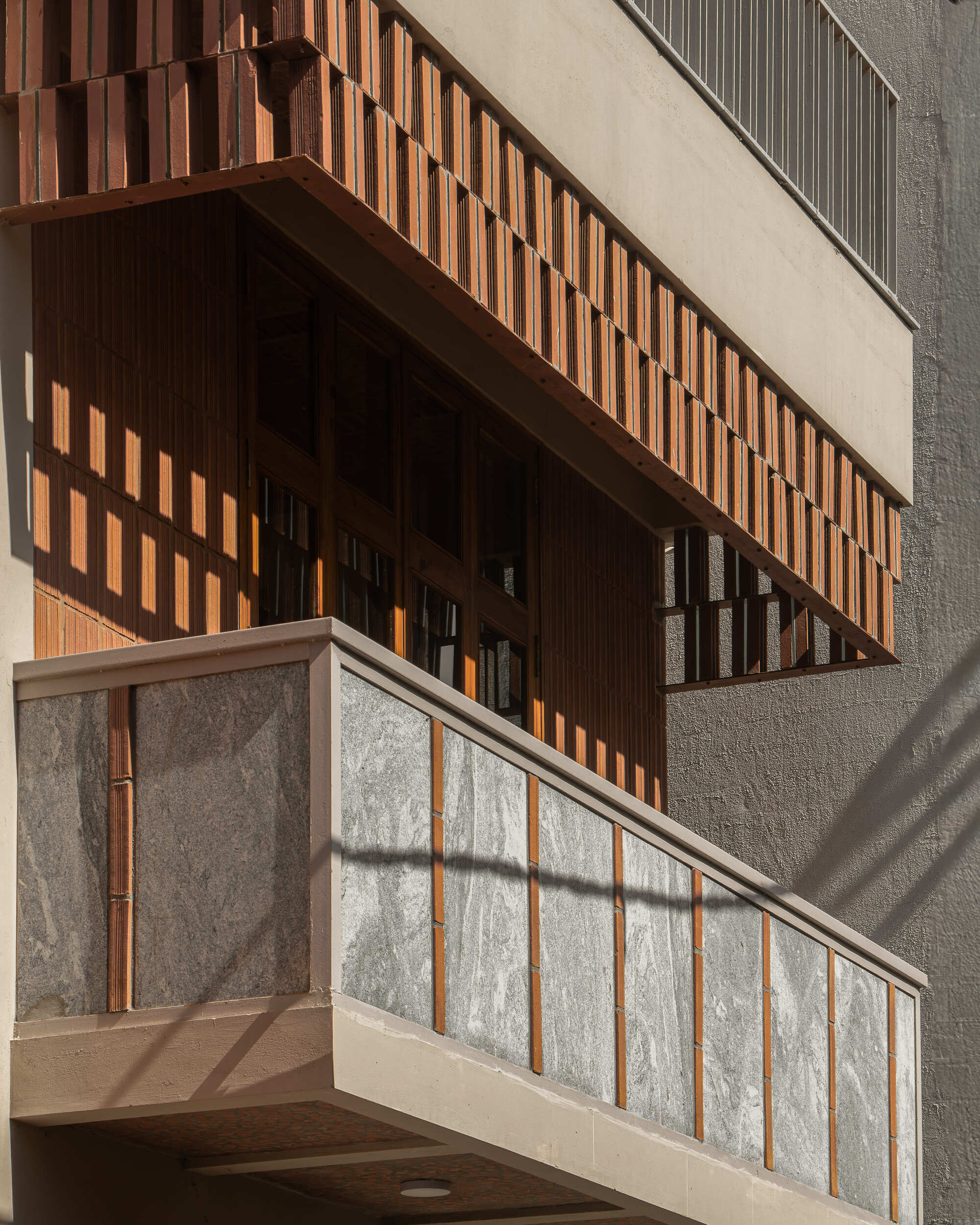 Facade detail showing timber slat balcony screen above textured stone panel railing in afternoon sunlight